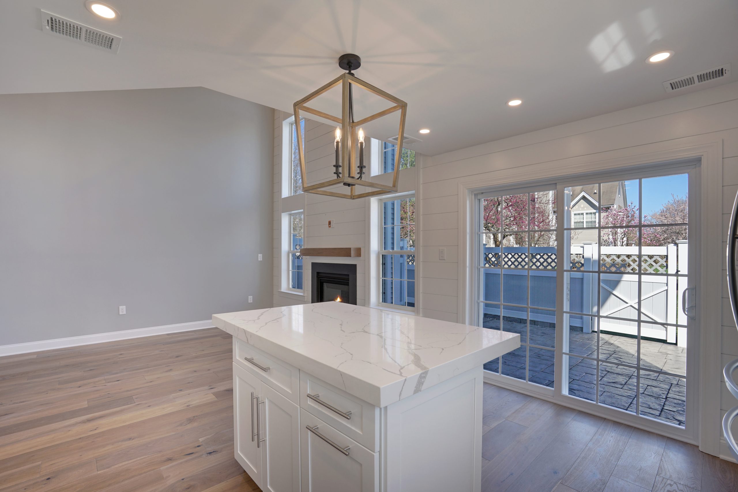 Modern kitchen with a white marble island, light wood floors, a large windowed door leading to a patio, a geometric chandelier, and a fireplace in the open living area.