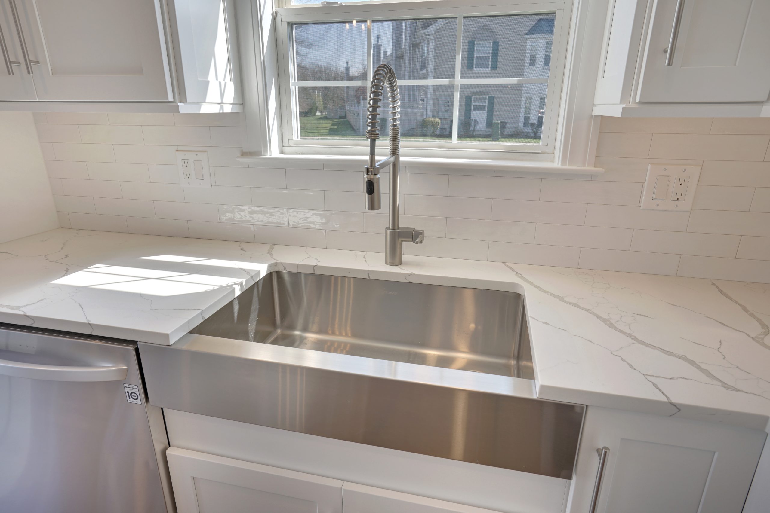 Modern kitchen with a large stainless steel farmhouse sink, white marble countertops, white subway tile backsplash, and a tall, flexible faucet beneath a window with a view of houses and green lawn outside.
