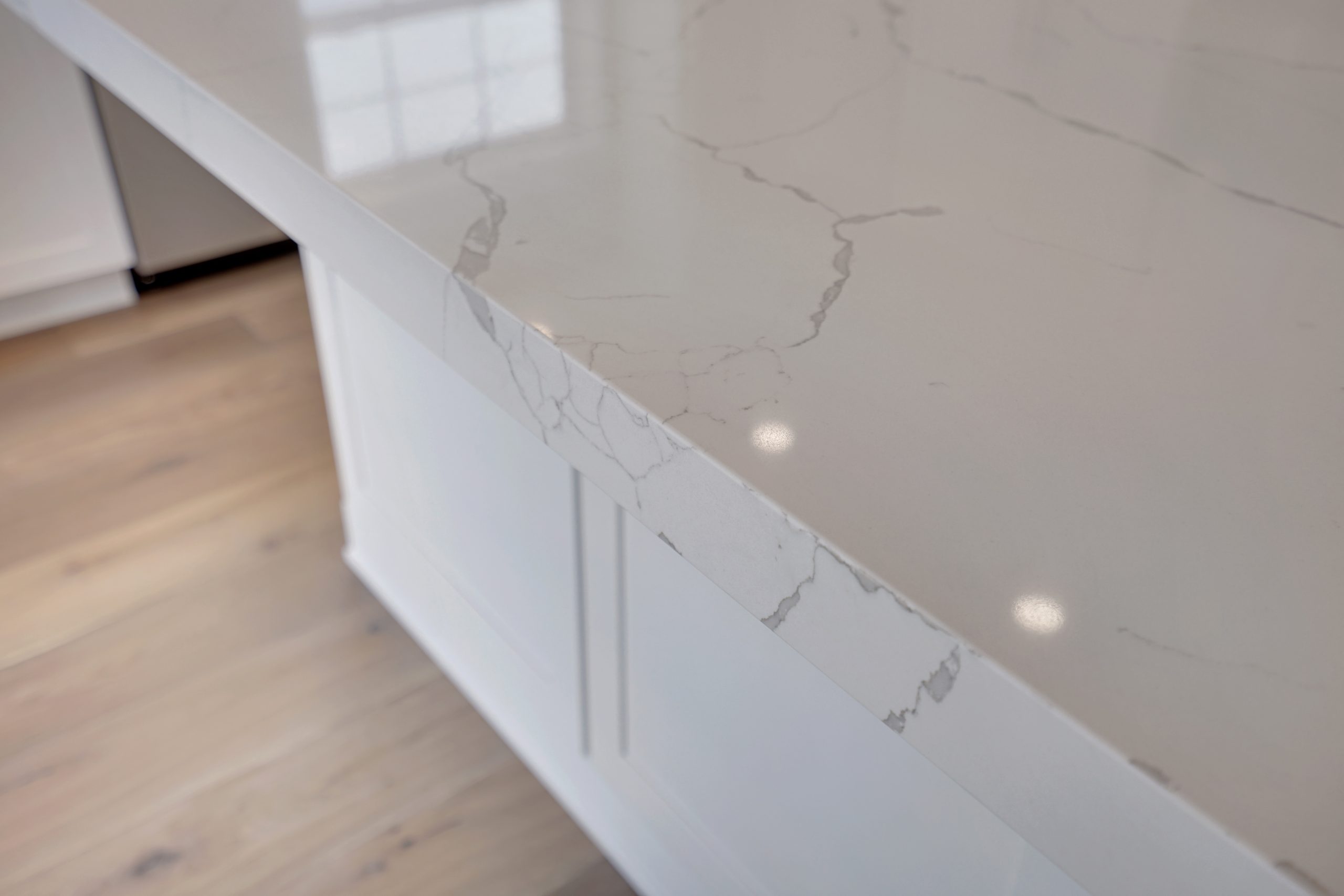Close-up of a light-colored marble or quartz countertop with subtle grey veining, polished surface, and white cabinetry below, reflecting ceiling lights on a light wood floor.