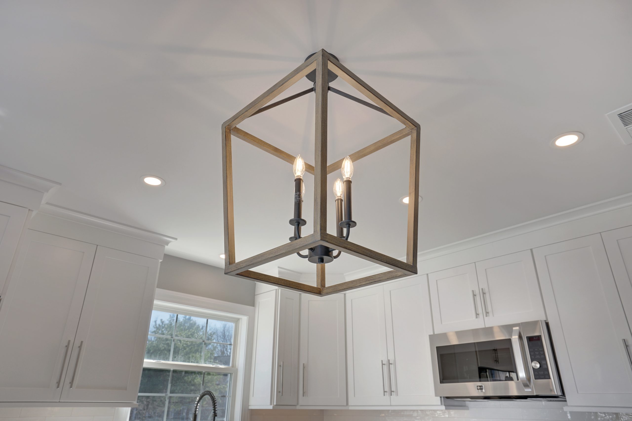 A modern kitchen light fixture with an open, geometric wooden frame and three exposed bulbs, hanging from a white ceiling with recessed lights and surrounded by white cabinets.
