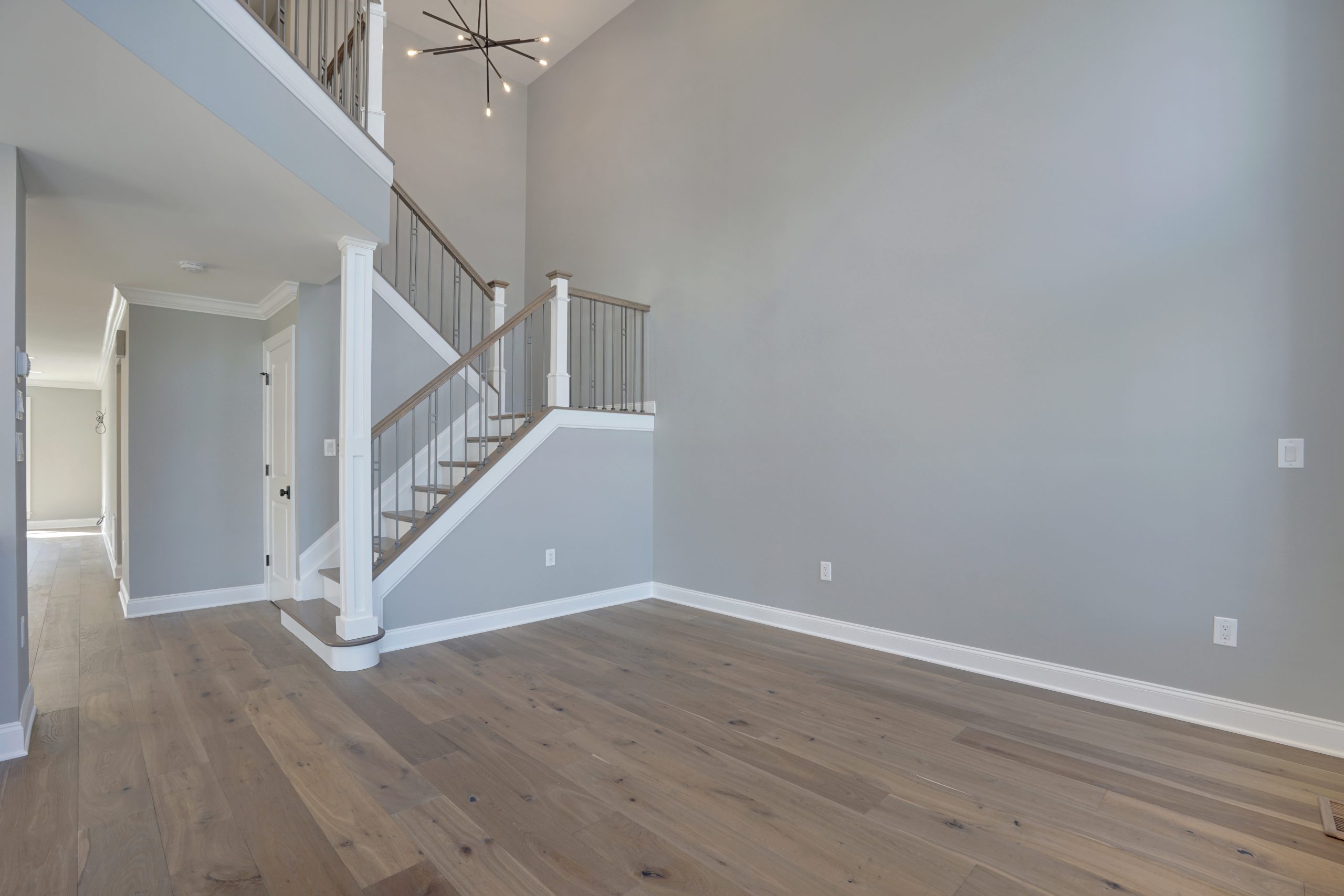 Modern, empty room with light wood floors, gray walls, a staircase with white and wood railings, and a contemporary chandelier hanging from a high ceiling. Natural light brightens the space.