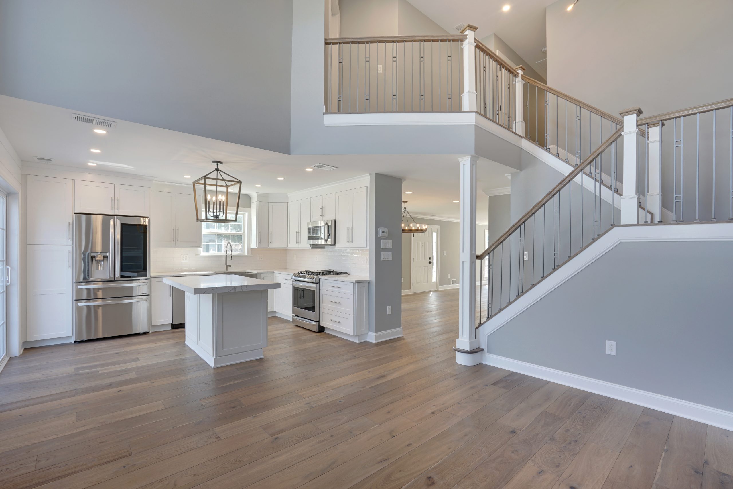 Modern open-concept kitchen and living space with white cabinetry, stainless steel appliances, an island, light wood flooring, and a staircase with gray accents and white railings leading to an upper level.
