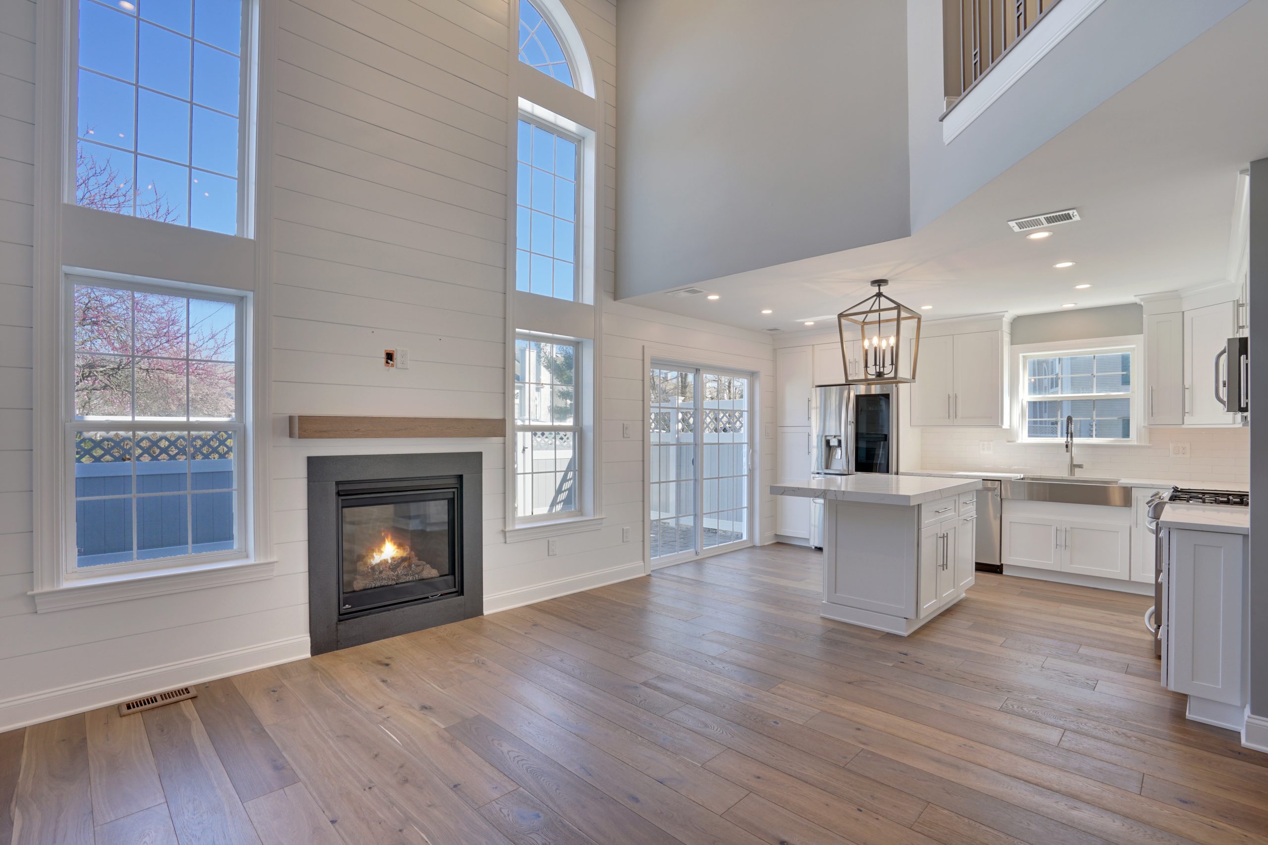 A modern, open-concept living area with large windows, a gas fireplace, light wood flooring, and a white kitchen with an island and pendant light, filled with natural sunlight.