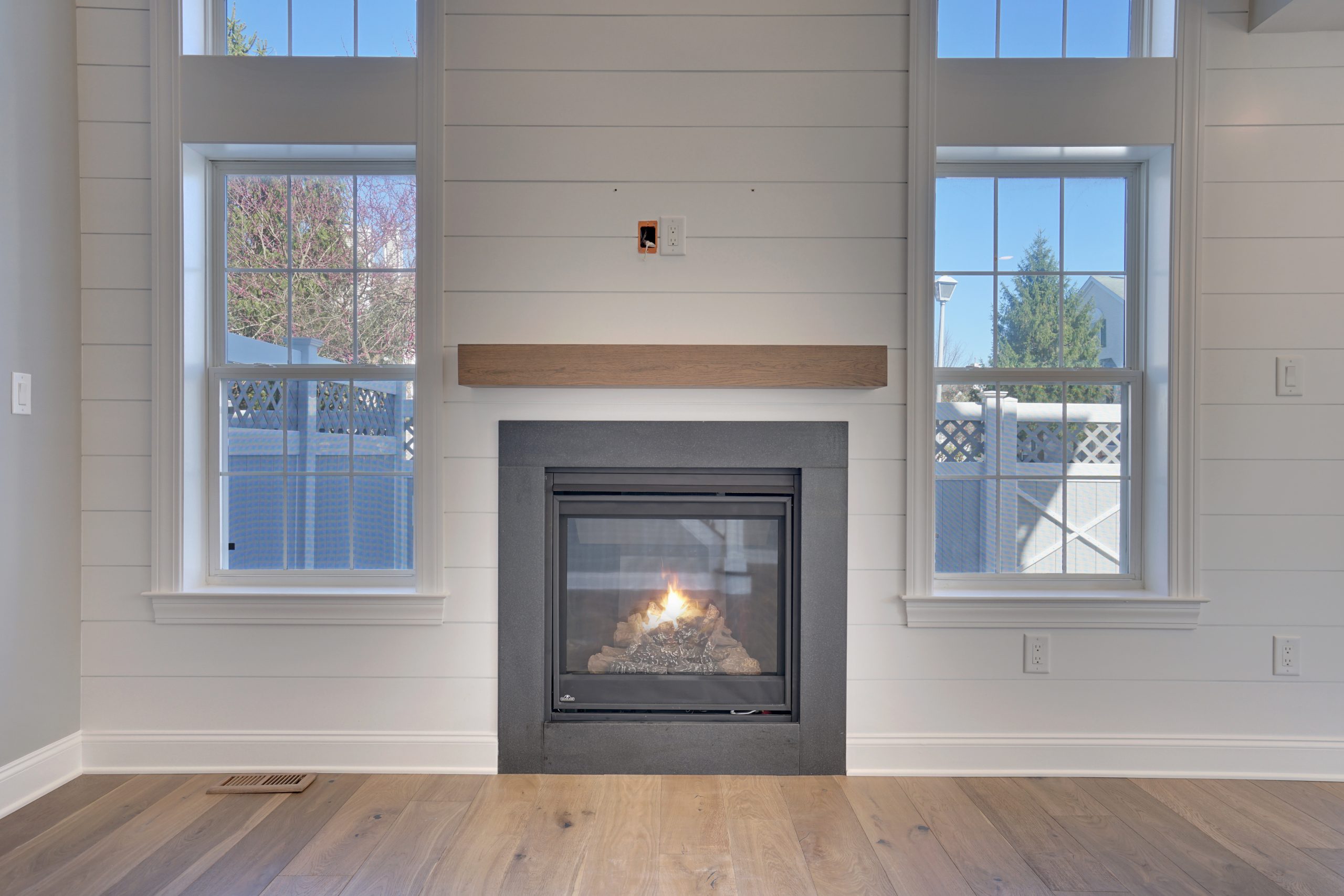 A modern living room with a gas fireplace centered between two large windows. The walls are white with shiplap paneling, and the floor has light wood planks. Outdoor trees are visible through the windows.