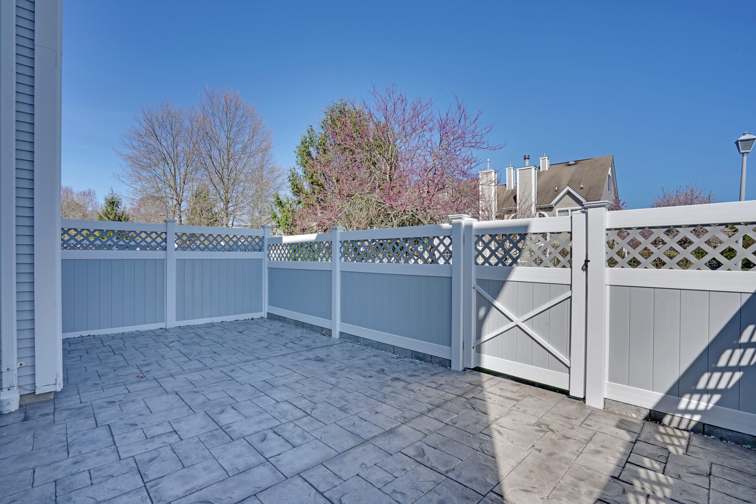 A small patio with light gray stamped concrete flooring, enclosed by a white and gray vinyl fence with a gate and lattice top, with trees and houses visible in the background under a clear blue sky.