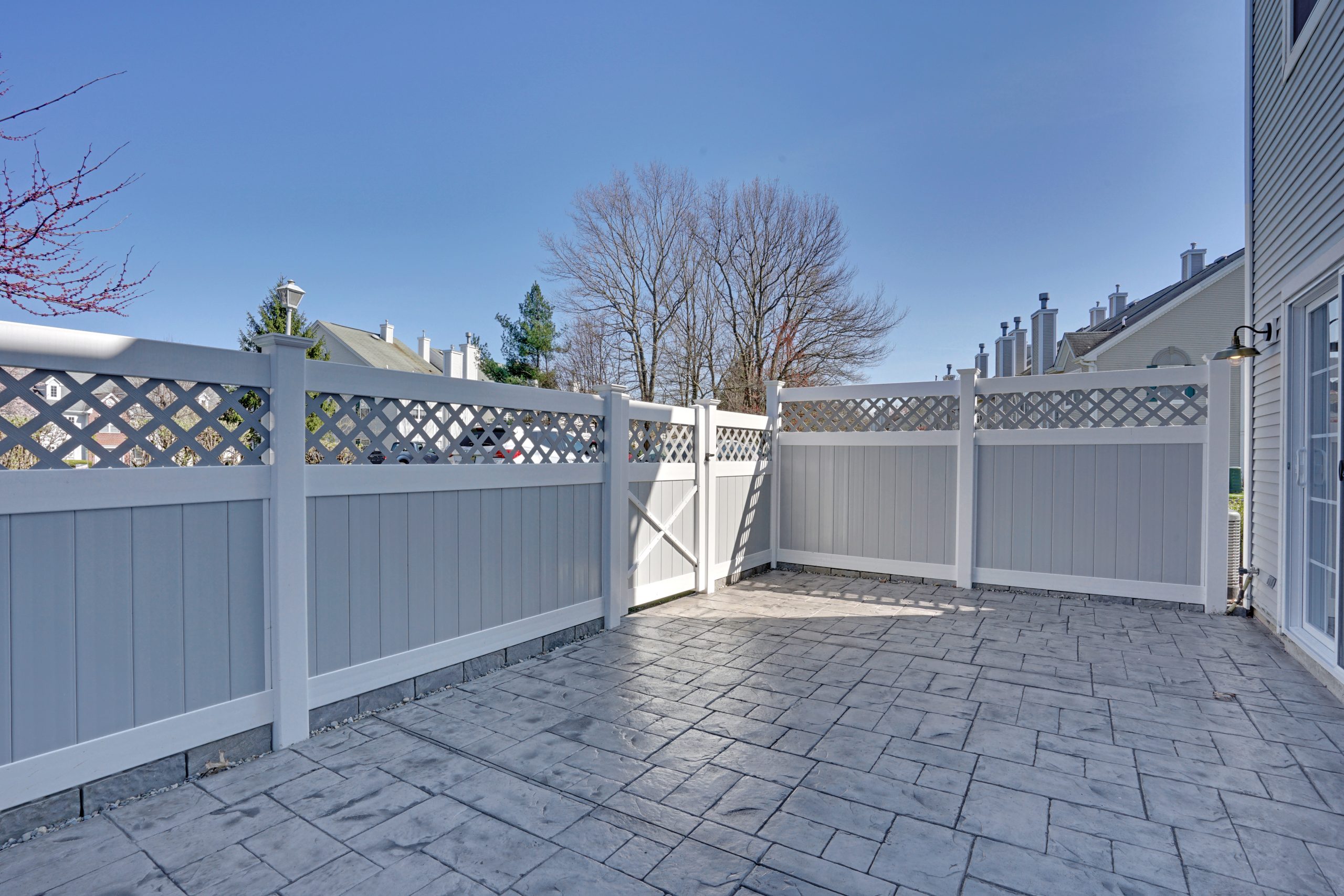 A clean, spacious patio with gray stamped concrete flooring, surrounded by a tall white and gray privacy fence with a lattice design at the top, under a clear blue sky.