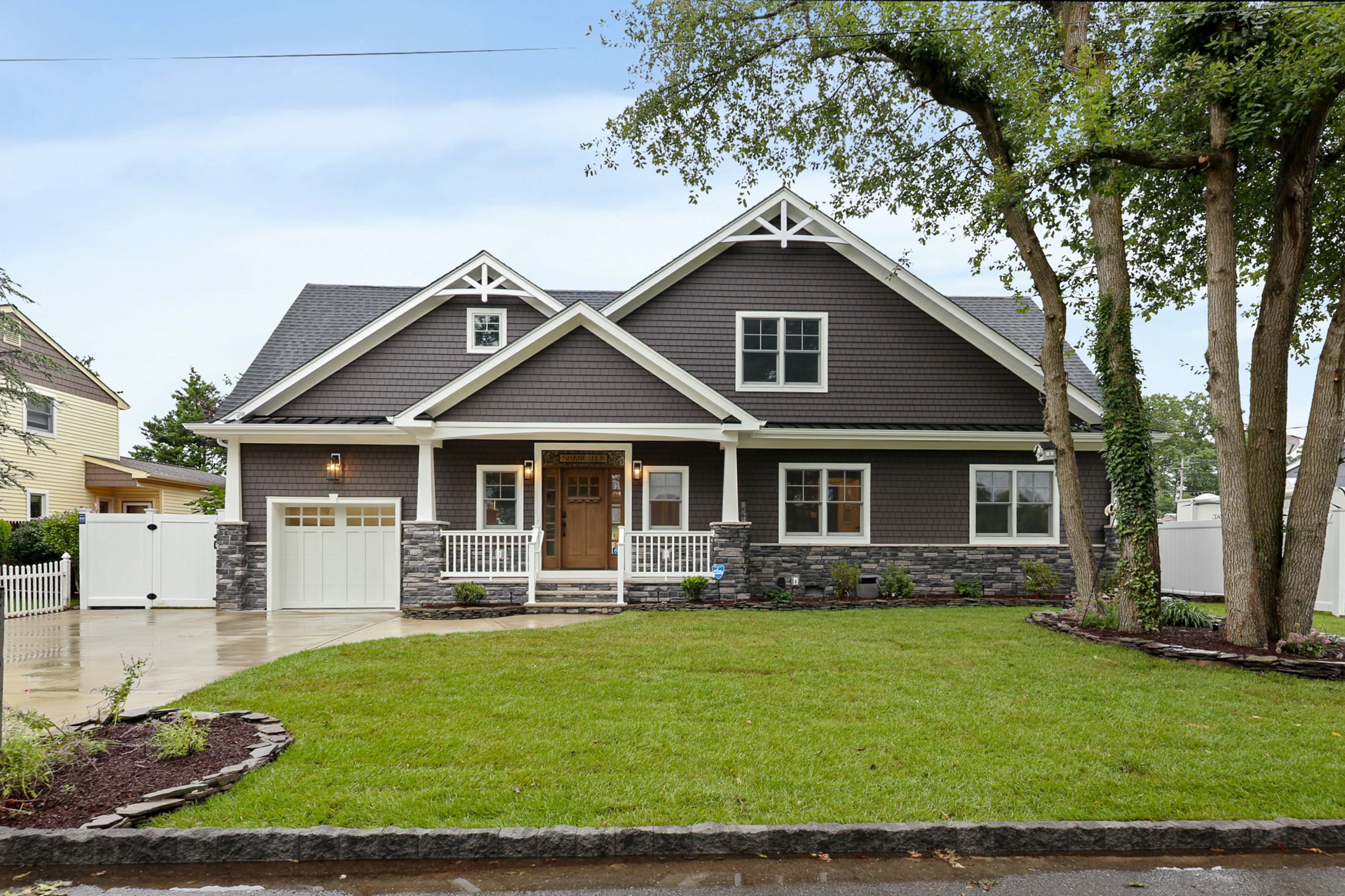 A two-story suburban house with brown siding, white trim, a covered front porch, stone accents, attached garage, and a neatly landscaped lawn with trees and shrubs.