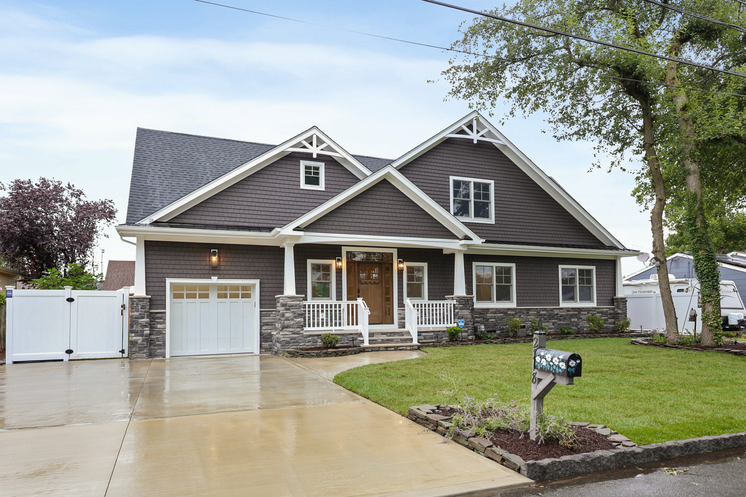 A modern two-story house with gray siding, stone accents, white trim, a covered front porch, and a single-car garage. The driveway and lawn are neatly maintained, and there are trees and mailboxes in front.