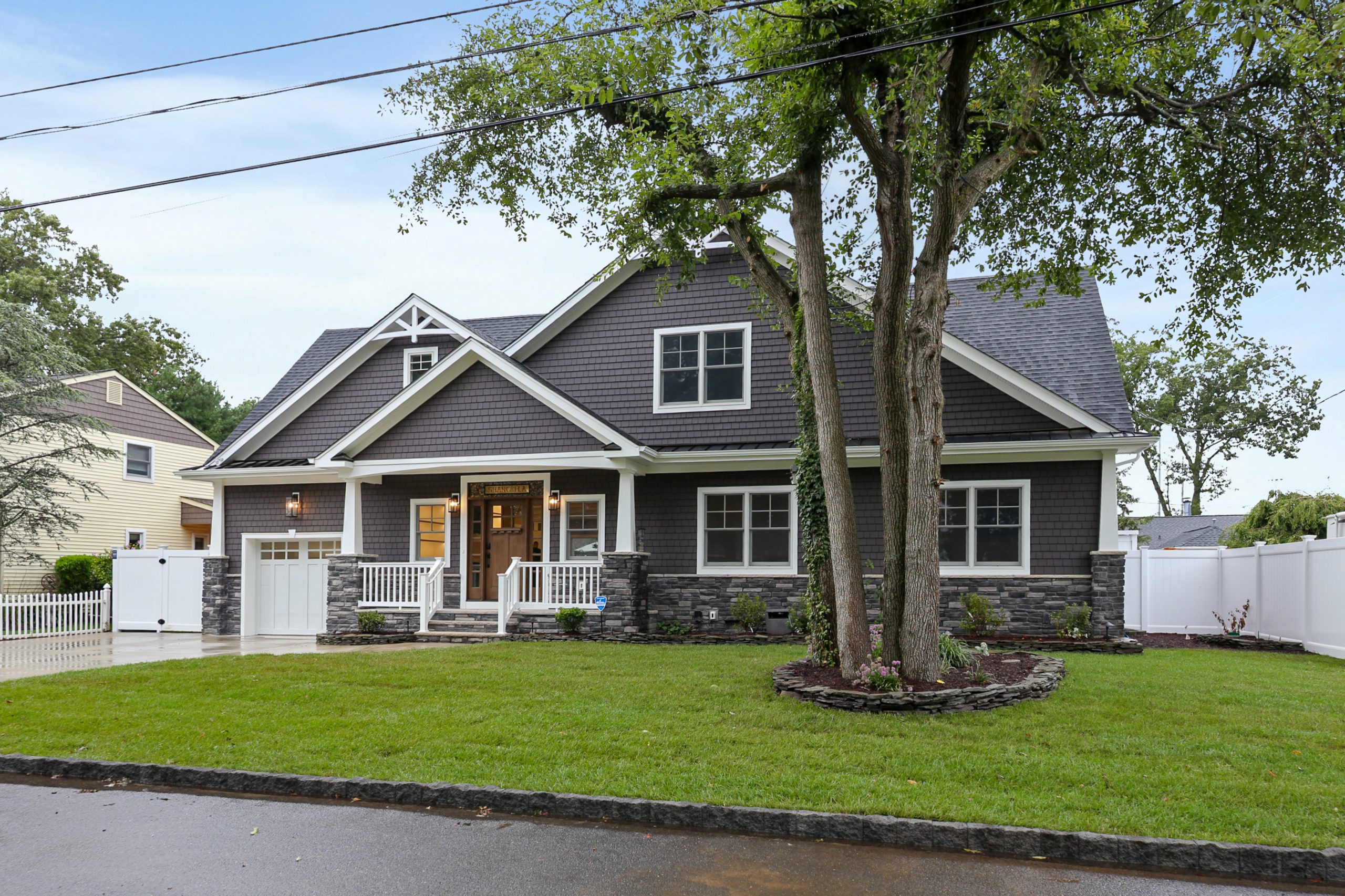 A modern two-story house with dark gray siding, stone accents, white trim, and a front porch. There is a neatly manicured lawn, trees, and a white fence surrounding the property.