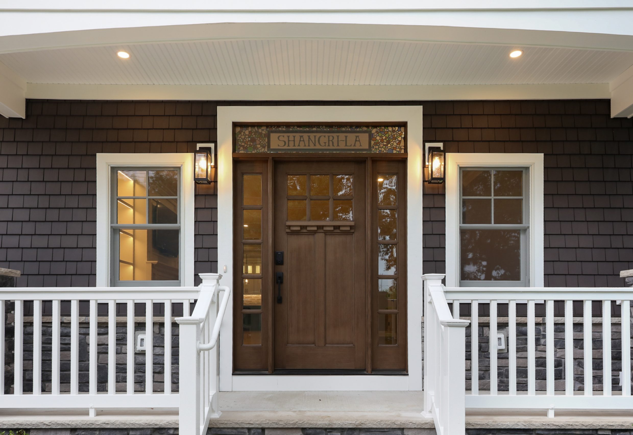 A wooden front door with glass panels is centered between two windows and lit by wall lanterns. Above the door is a decorative sign that reads SHANGRI-LA. White railing and steps lead to the entrance.
