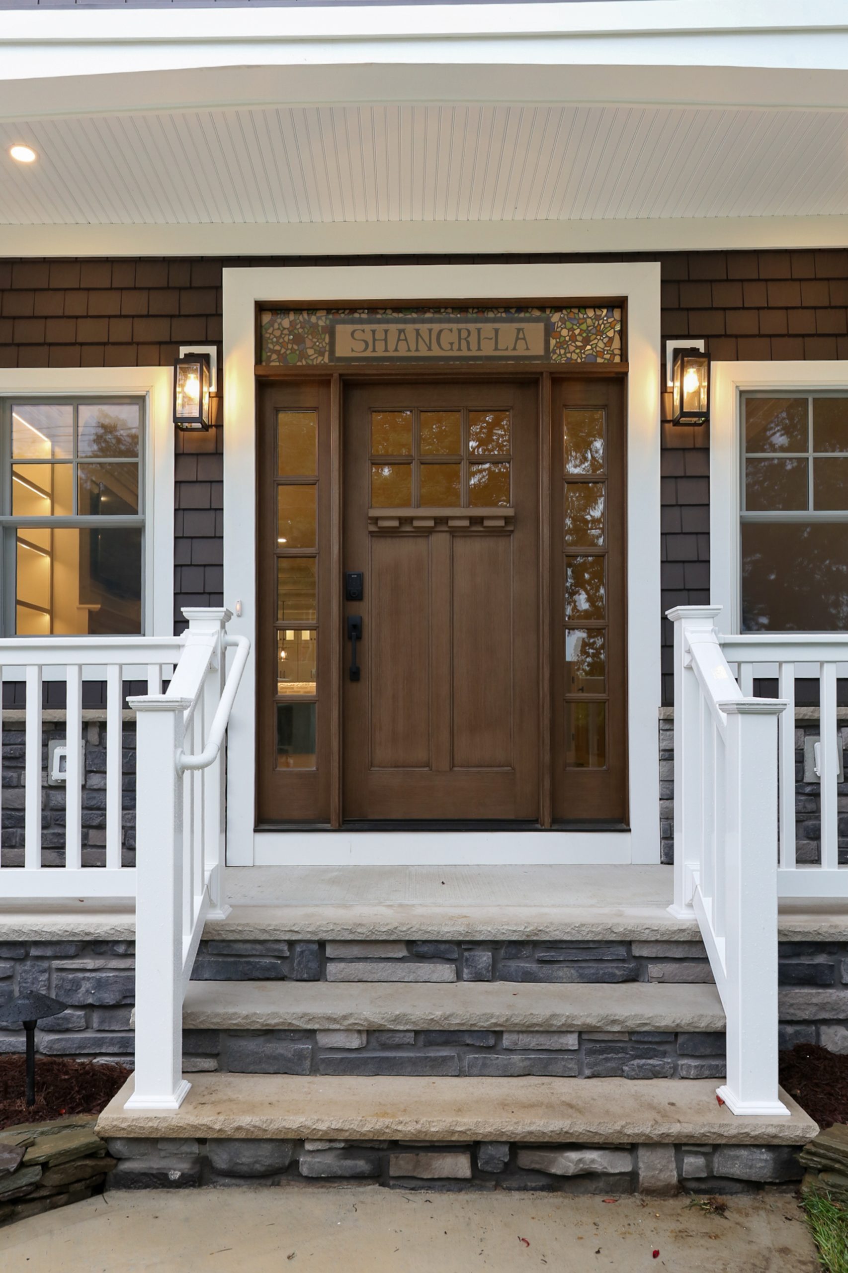 A wooden front door with glass panels and a sign reading SHANGRI-LA above it, flanked by two lit lanterns. White railings and stone steps lead up to the entrance of a house with gray siding.