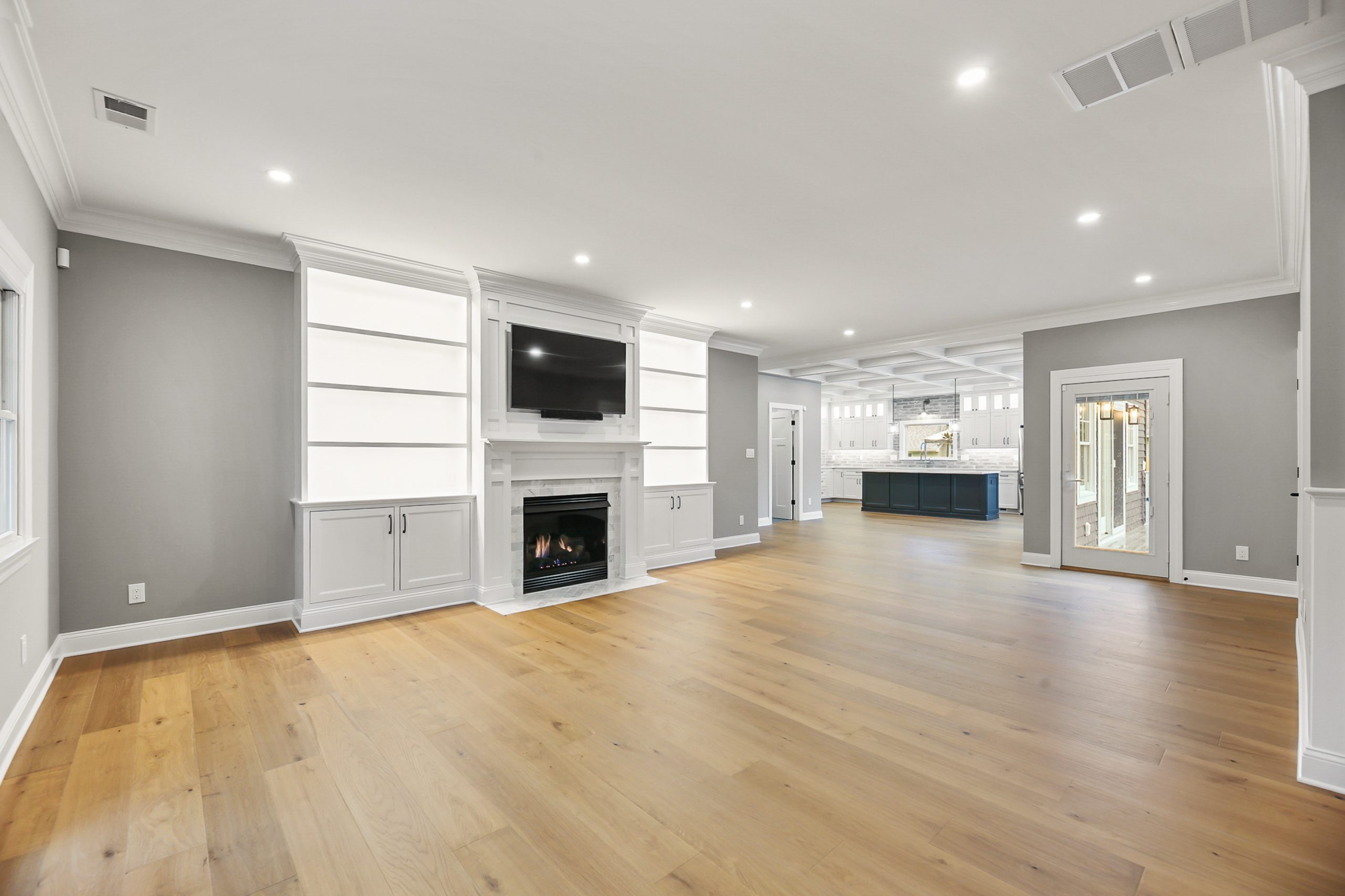 Spacious, modern living room with light wood flooring, gray and white walls, built-in shelves, a fireplace, and a mounted TV. The room opens to a kitchen area and has large windows and glass doors letting in natural light.