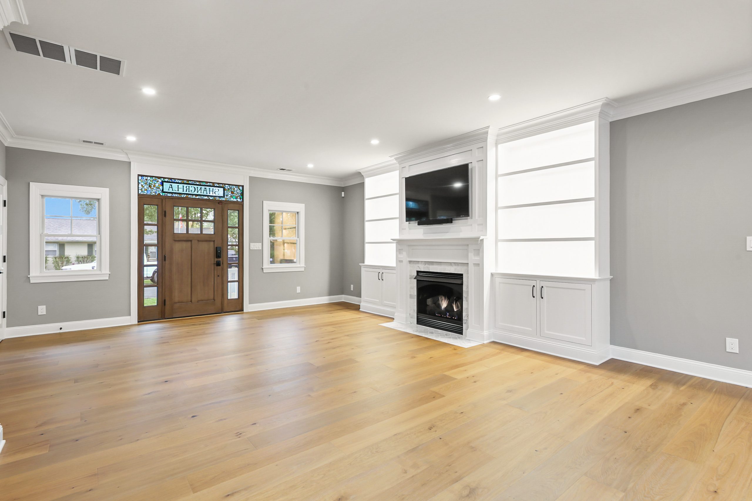 Bright living room with light wood floors, gray walls, white trim, built-in shelves, a fireplace with a TV above, and a wooden front door featuring stained glass and sidelights. Multiple windows let in natural light.