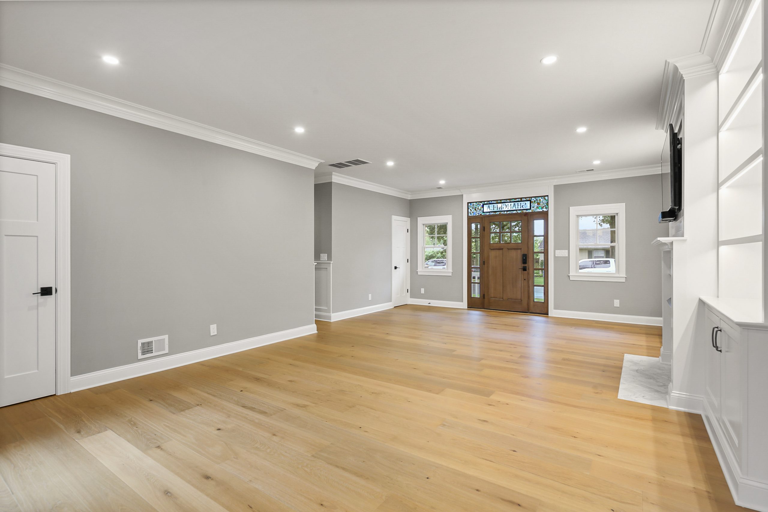 Bright, modern living room with light wood flooring, gray walls, white trim, and built-in shelves. Features recessed lighting, a wooden front door with glass panels, and windows allowing natural light into the space.