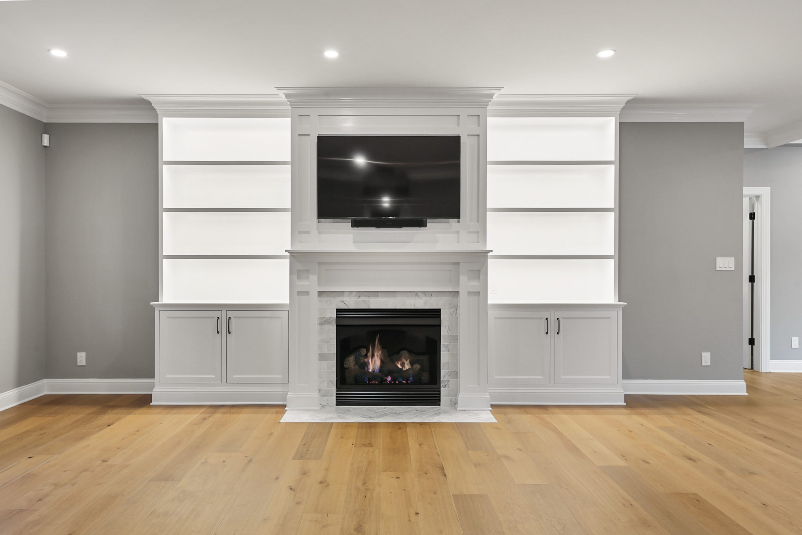 A modern living room with light wood floors, gray walls, built-in white shelves, a fireplace below a flat-screen TV, and recessed ceiling lights. The shelves are empty, and the room is uncluttered and bright.