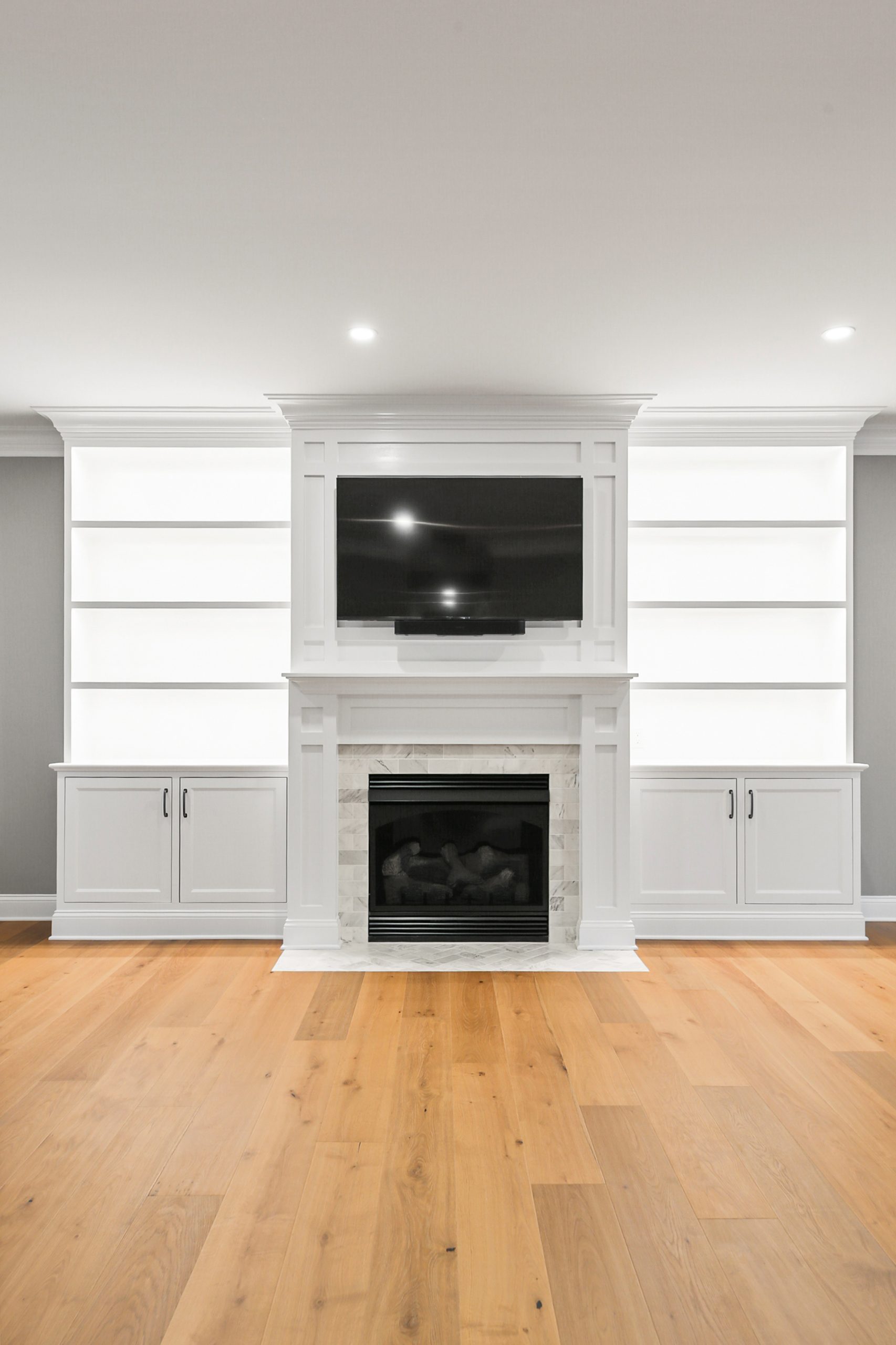 A modern living room with a built-in white shelving unit, a flat-screen TV mounted above a white marble fireplace, closed cabinets below, and light wood floors. The shelves are empty and the space is brightly lit.