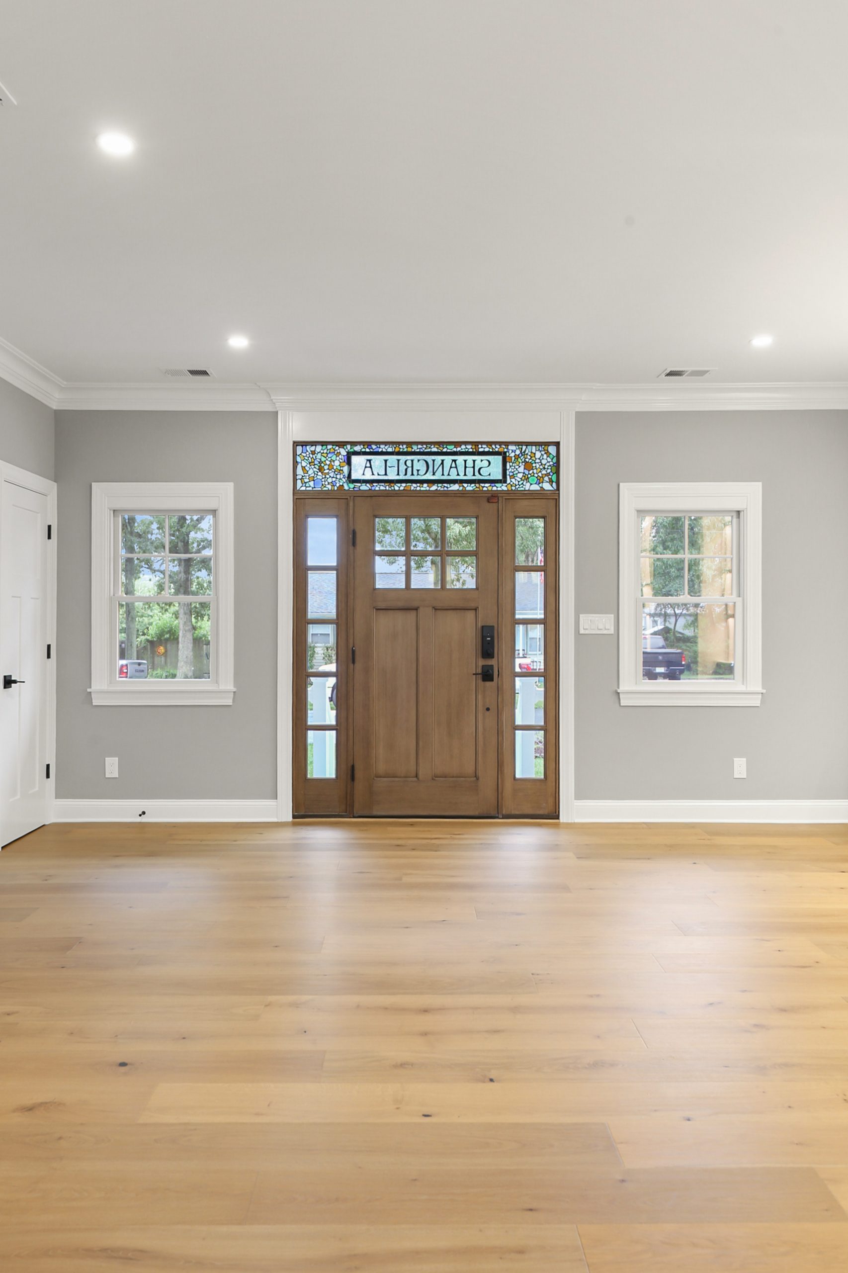 A bright entryway with light wood floors, gray walls, and a wooden front door featuring stained glass above it, reading “SHANGRILA.” Two windows flank the door, and recessed lights illuminate the space.