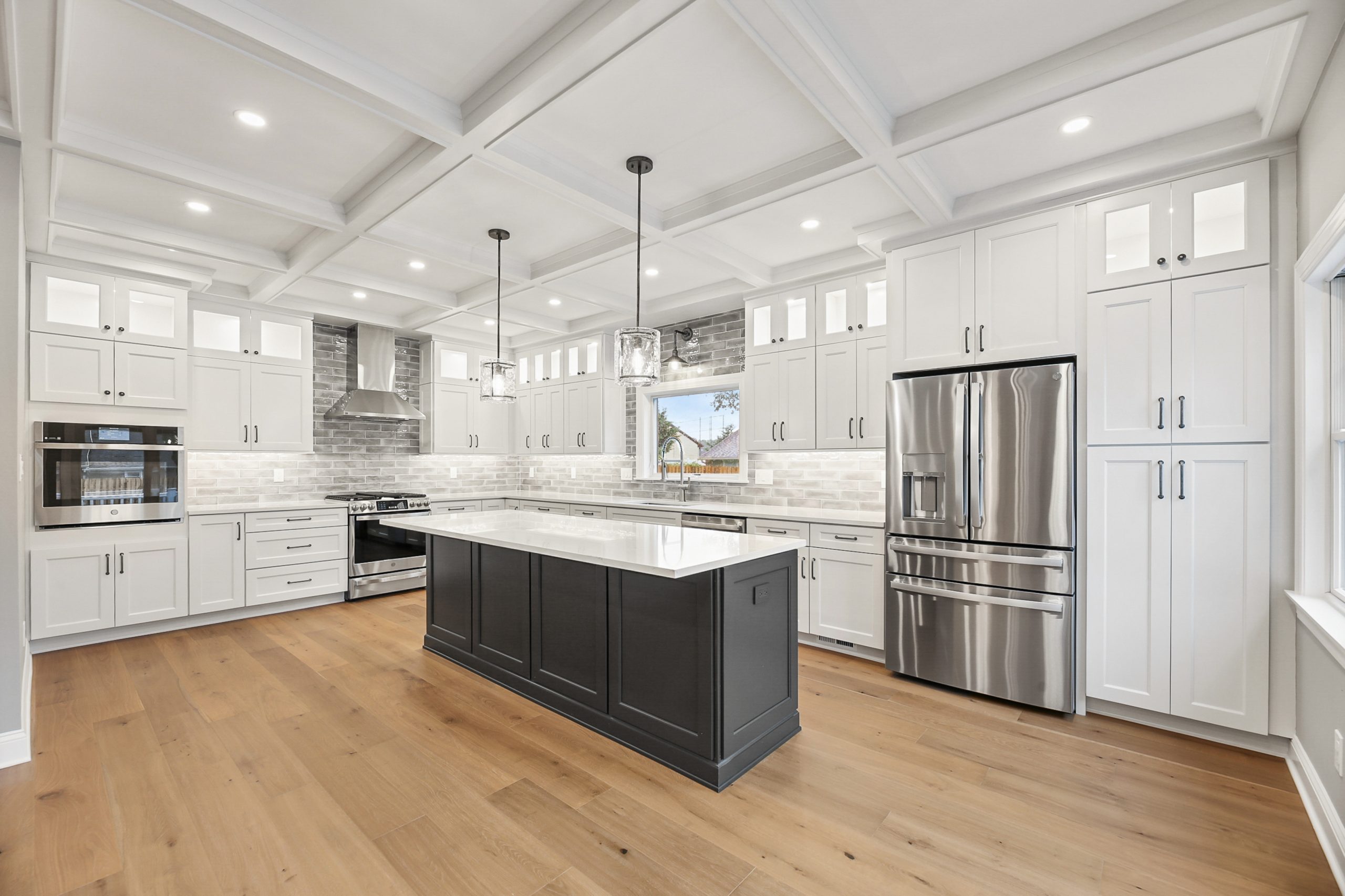 Modern kitchen with white cabinets, stainless steel appliances, a large dark gray island, pendant lights, light wood flooring, and a coffered ceiling. Subway tile backsplash and many windows create a bright, airy atmosphere.