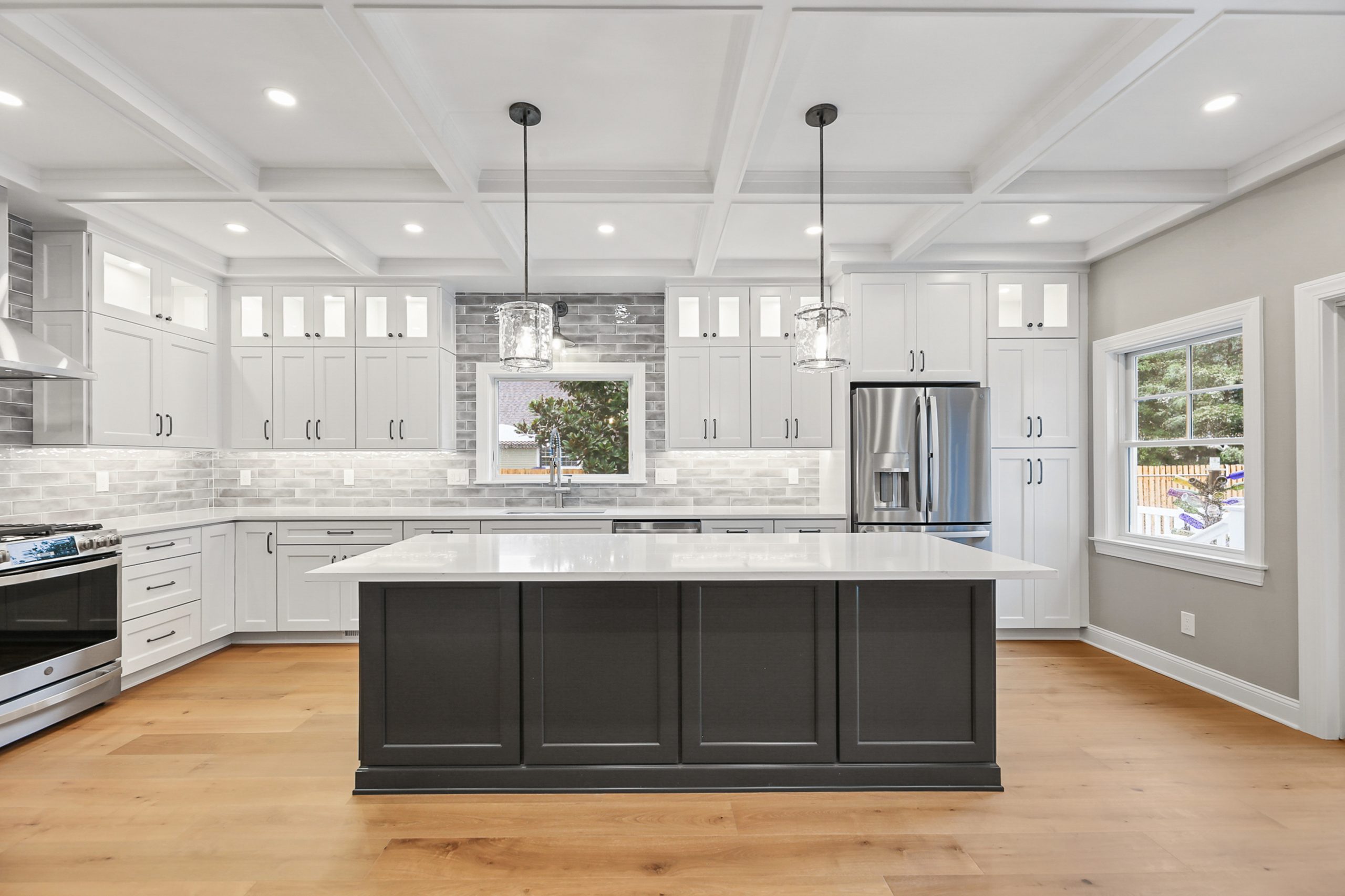 Modern kitchen with white cabinets, stainless steel appliances, a large dark island with a white countertop, pendant lights overhead, and light wood flooring. Large windows let in natural light.