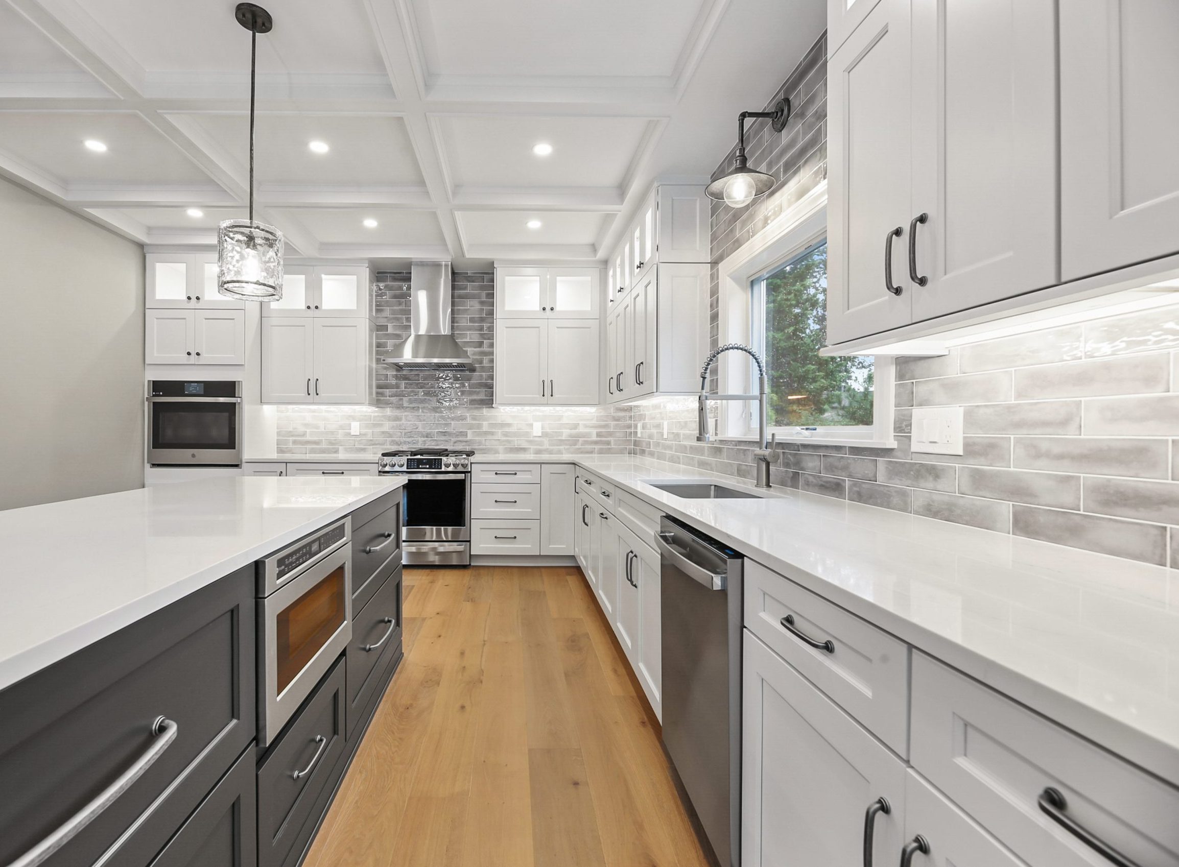 Modern kitchen with white cabinetry, gray tile backsplash, stainless steel appliances, large island with built-in microwave, pendant lighting, farmhouse sink, and natural wood flooring. Bright and spacious with coffered ceiling.
