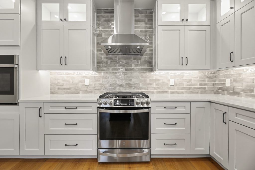 Modern kitchen with white cabinets, stainless steel gas stove and range hood, gray subway tile backsplash, under-cabinet lighting, and light wood flooring. Countertops are white and the overall look is clean and contemporary.