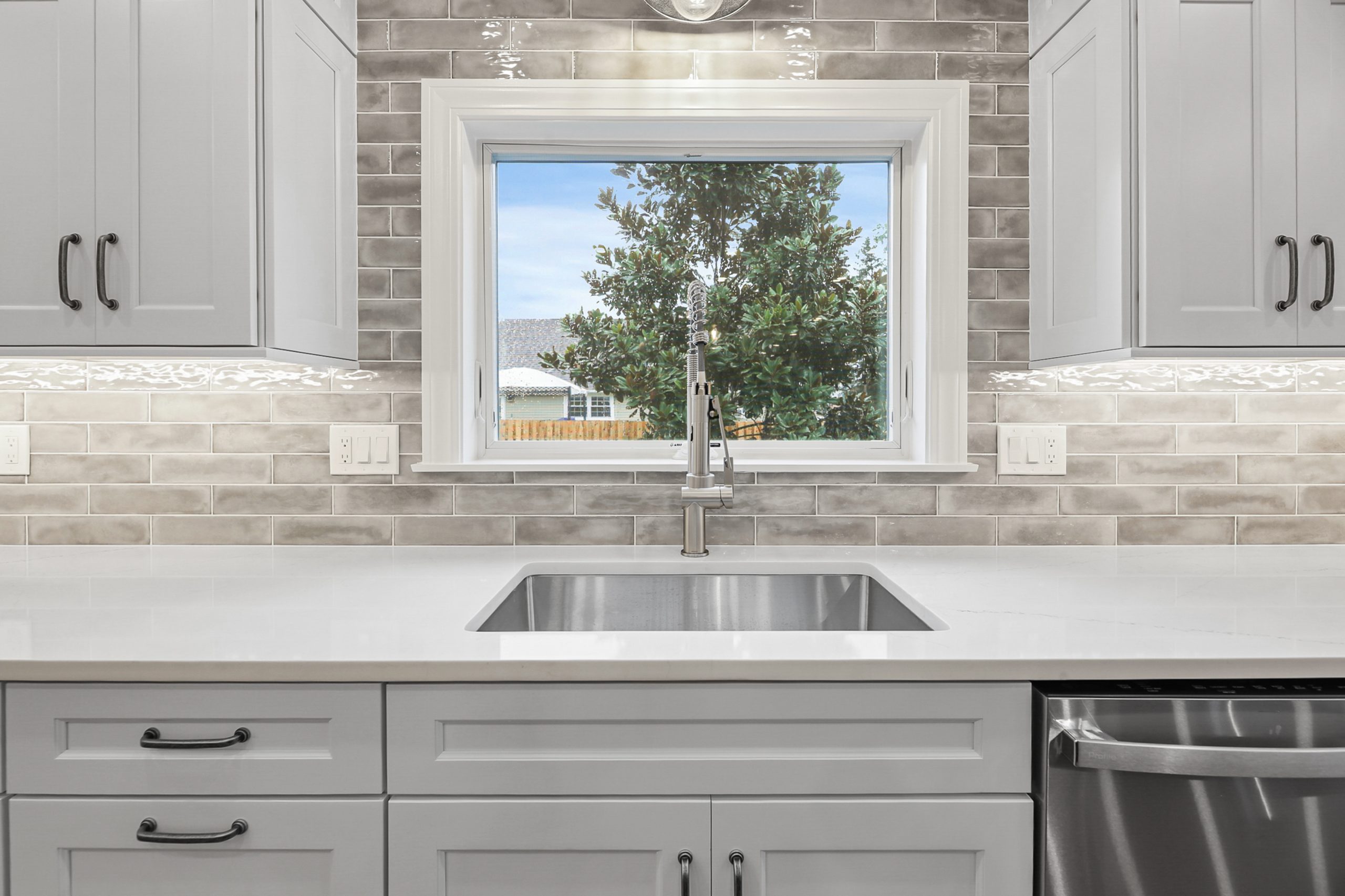 A modern kitchen sink with a stainless steel faucet sits beneath a window. White cabinets, gray subway tile backsplash, and white countertops create a clean, bright look. A tree is visible outside the window.