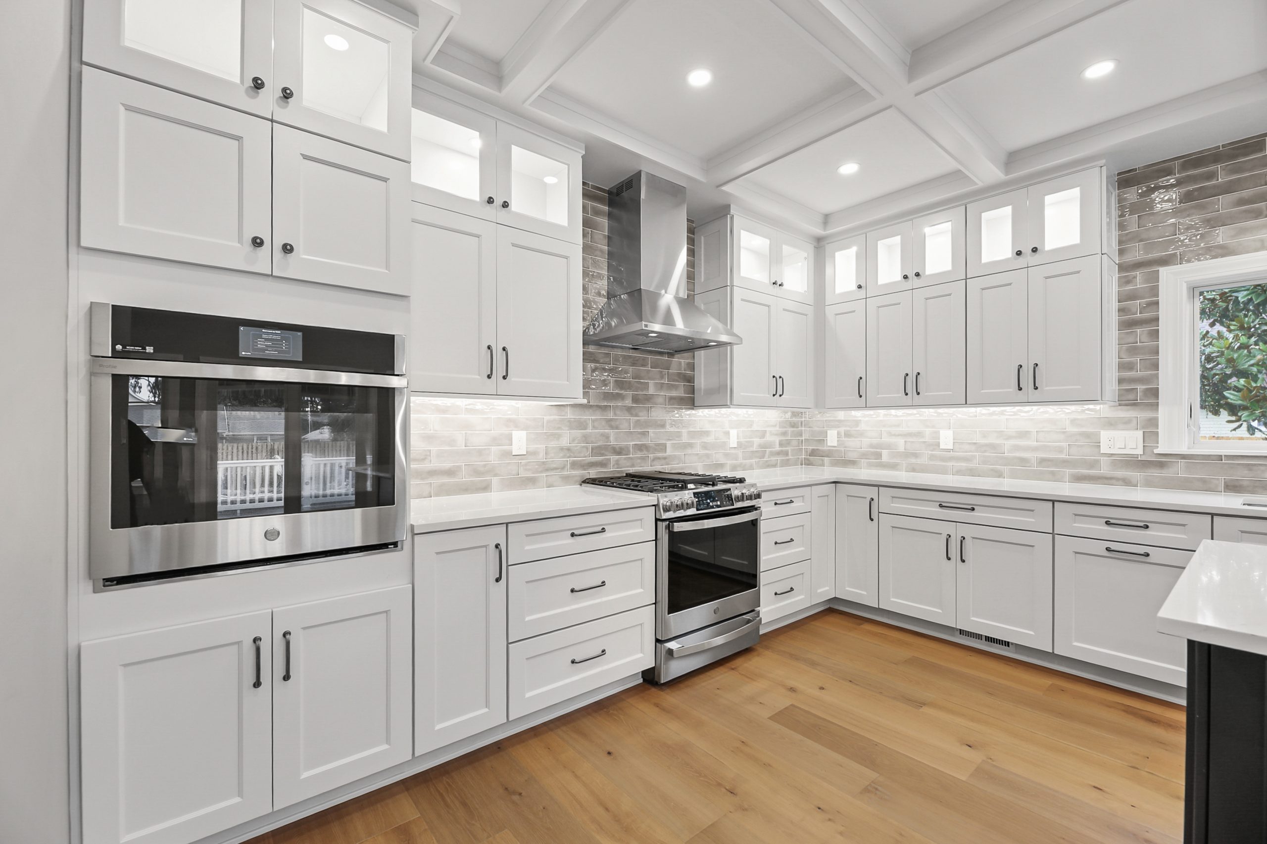 Modern kitchen with white cabinets, stainless steel appliances, a built-in oven, gas stove with hood, light wood flooring, gray subway tile backsplash, and recessed lighting.