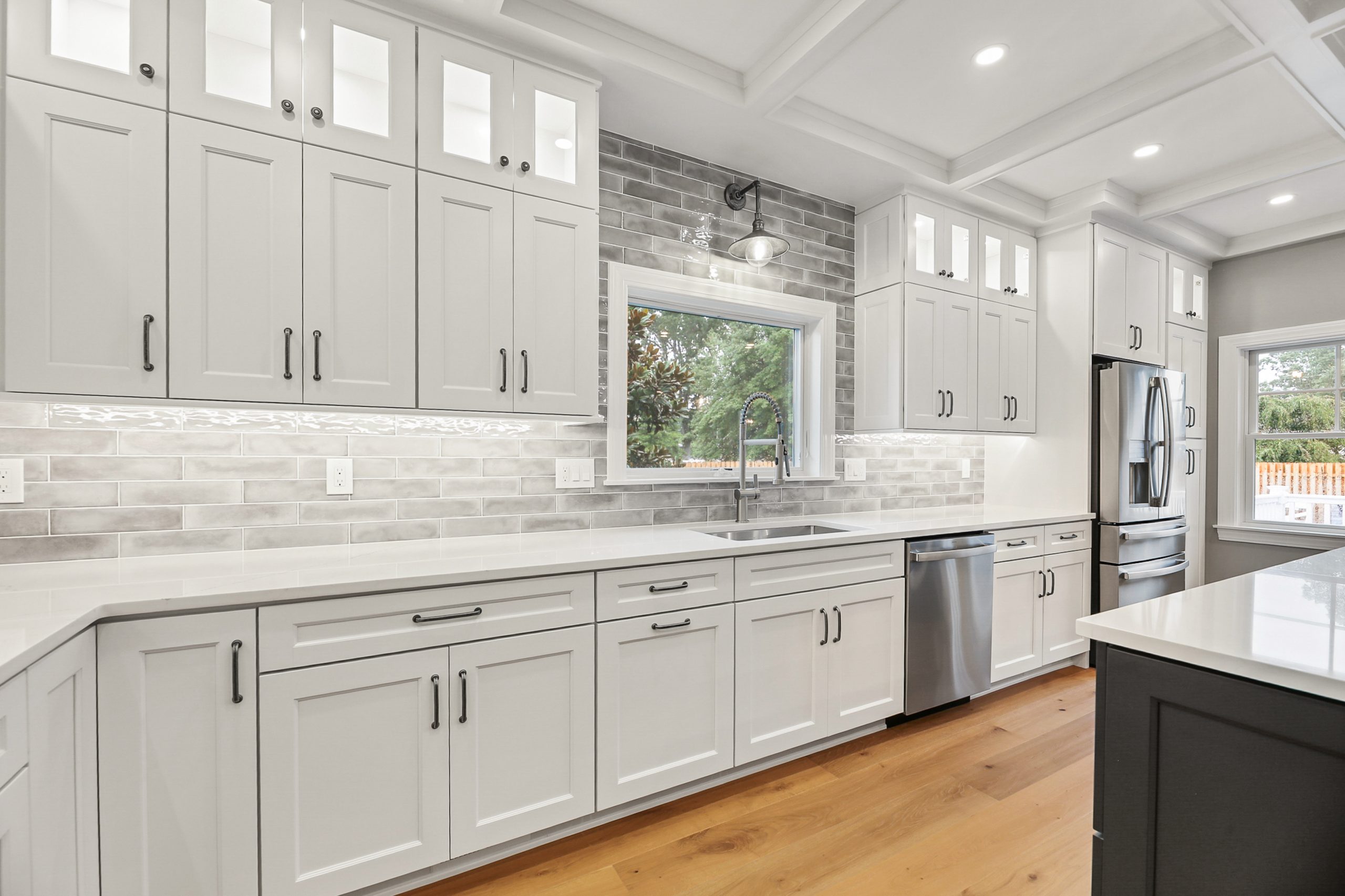 Modern kitchen with white cabinets, gray subway tile backsplash, stainless steel appliances, large windows, a farmhouse sink, and wood flooring. Ample counter space and recessed lighting create a bright, clean look.