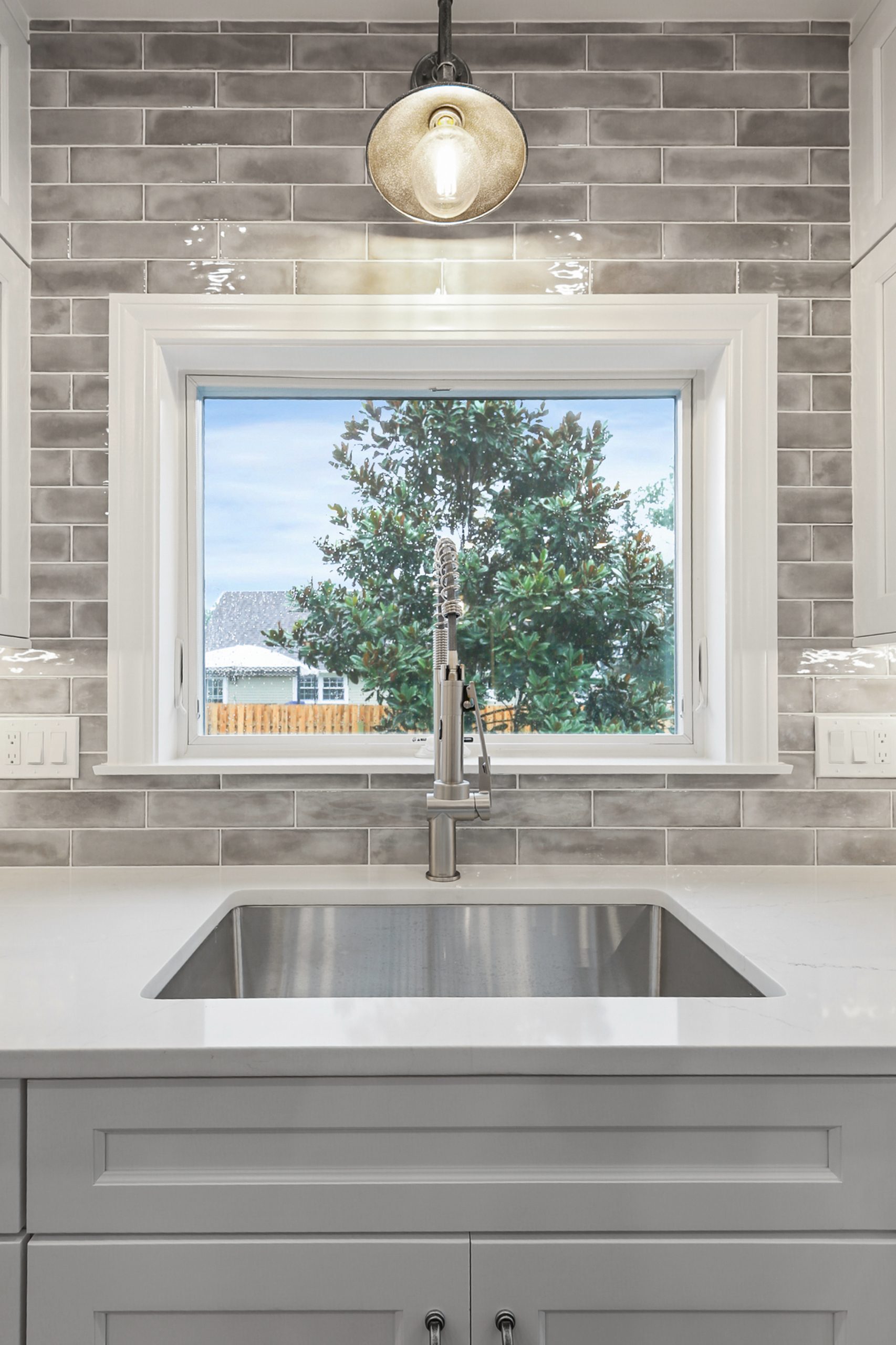 Modern kitchen sink with a sleek faucet is set under a large window. Grey subway tiles line the backsplash, and a pendant light hangs above. Outside, a tree and part of a fence are visible through the window.