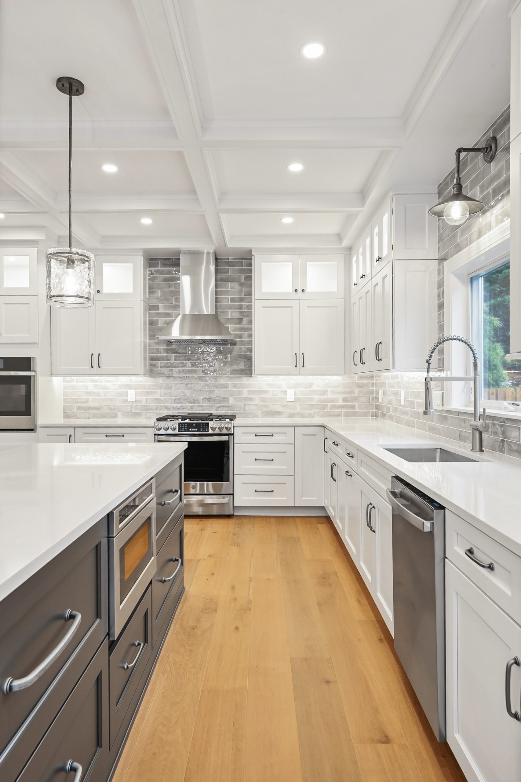 Modern kitchen with white cabinets, stainless steel appliances, light gray subway tile backsplash, large island with gray base, built-in oven, pendant lighting, and light wood flooring. Bright, clean, and spacious design.
