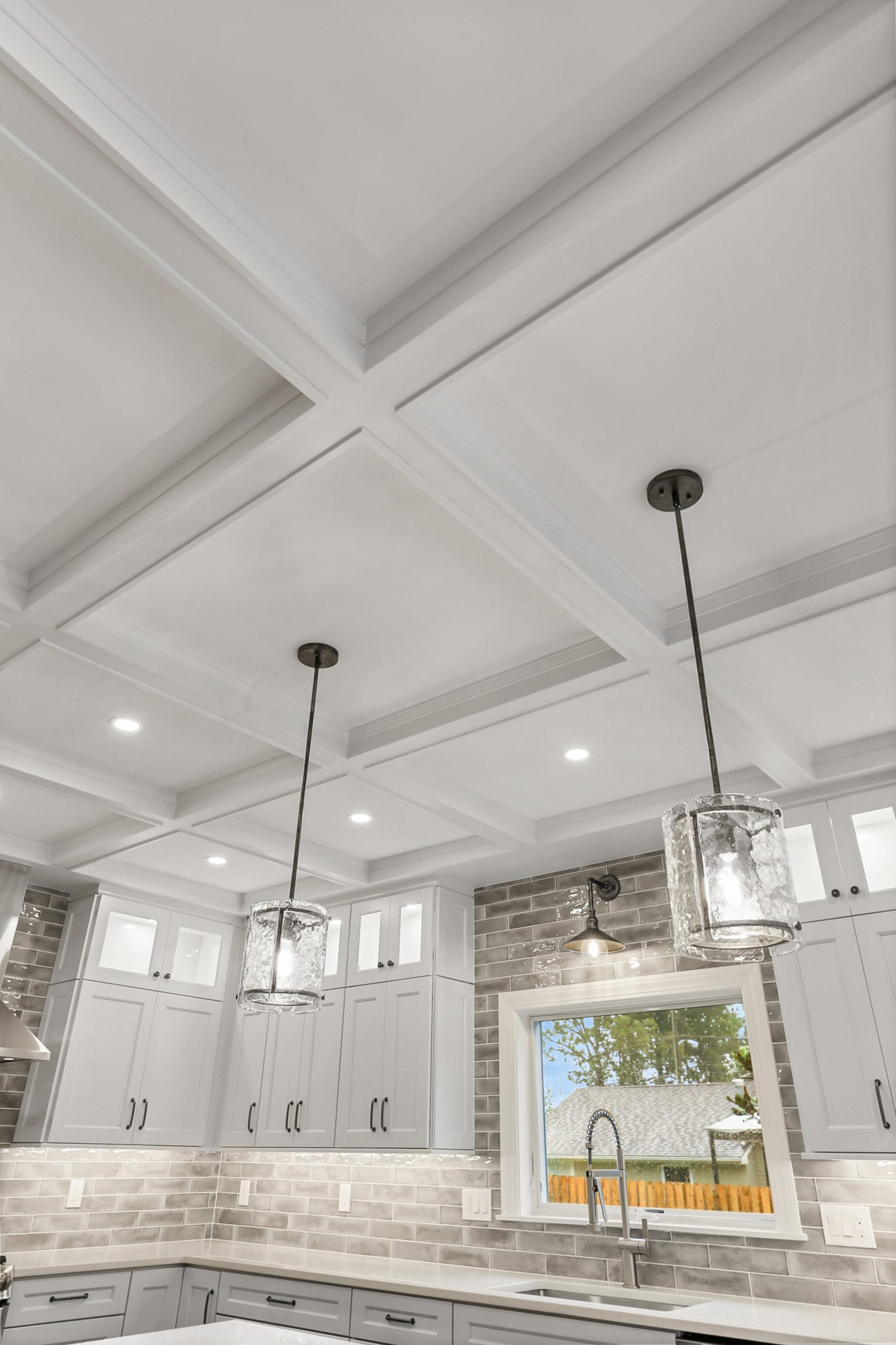 Modern kitchen with a coffered ceiling, two pendant lights over the counter, gray cabinets, brick-patterned backsplash, and a window above the sink letting in natural light.