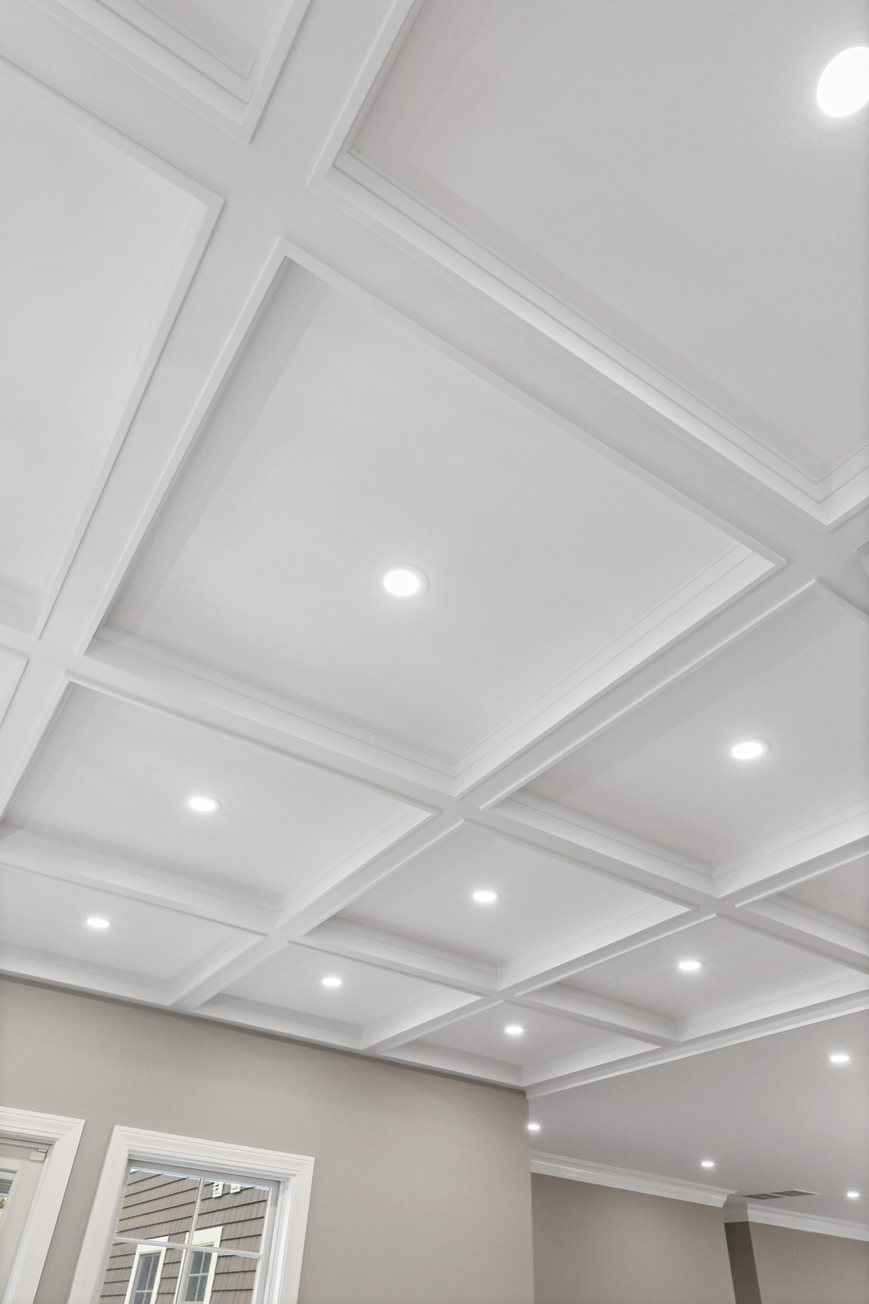 White coffered ceiling with recessed lights in a modern, neutral-colored room. Natural light enters through windows at the bottom left corner of the image.