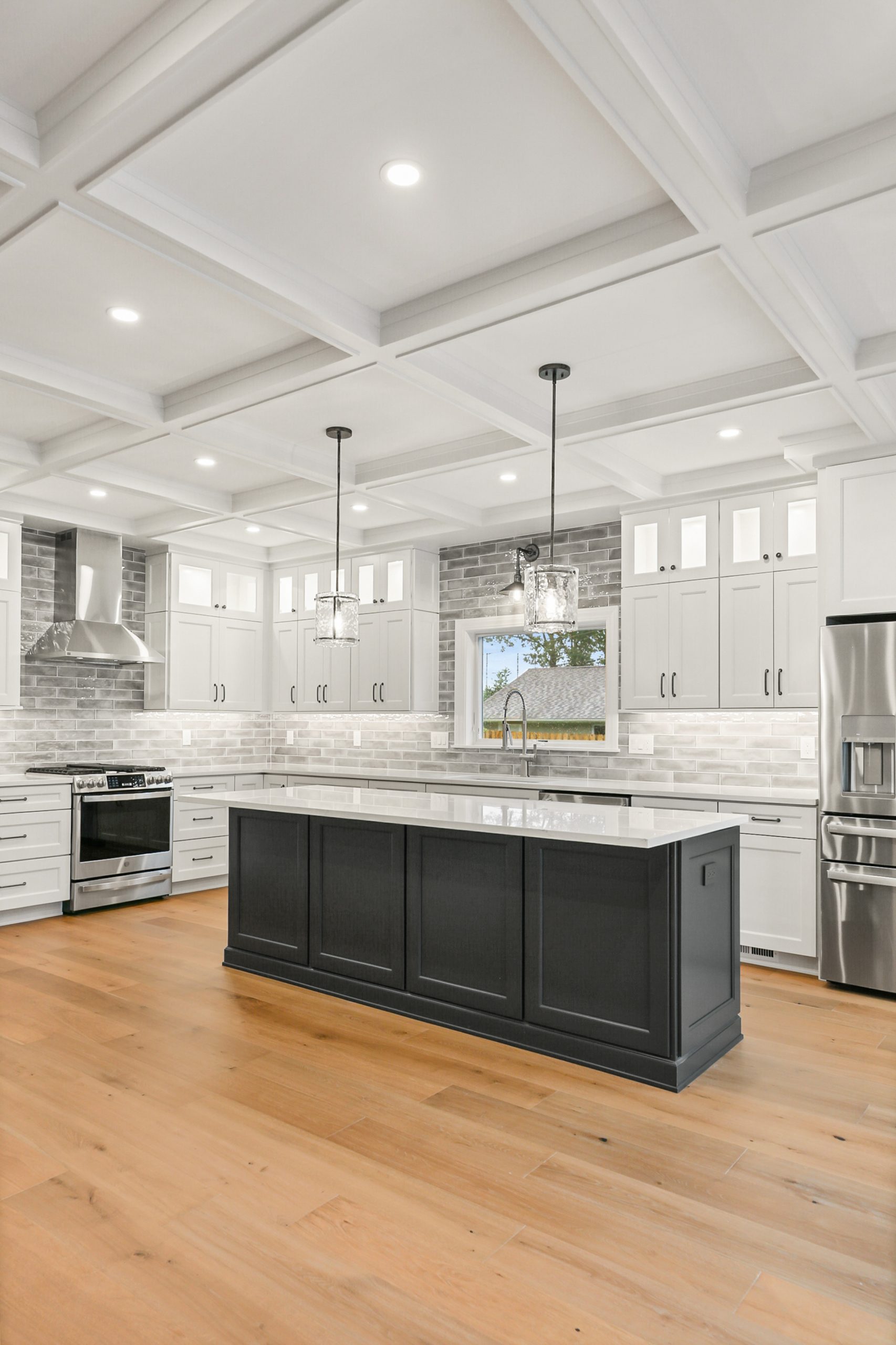 Modern kitchen with a large black island, stainless steel appliances, white cabinets, a brick backsplash, pendant lights, and light wood flooring. The space is bright with recessed ceiling lights and a window above the sink.