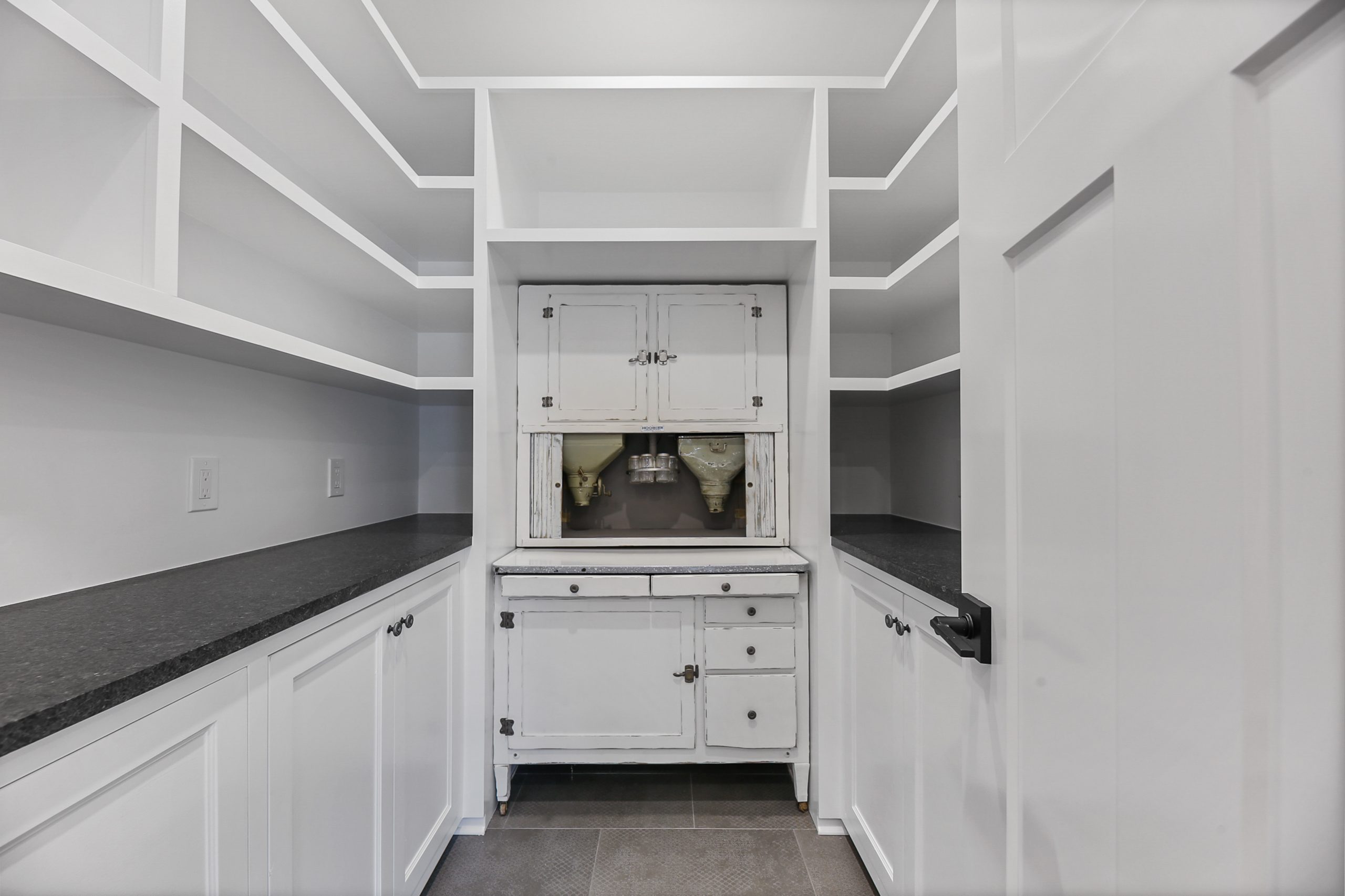 A white pantry with open shelves, black countertops, and a vintage white cabinet in the center. The room has clean lines, a gray floor, and a white paneled door on the right.
