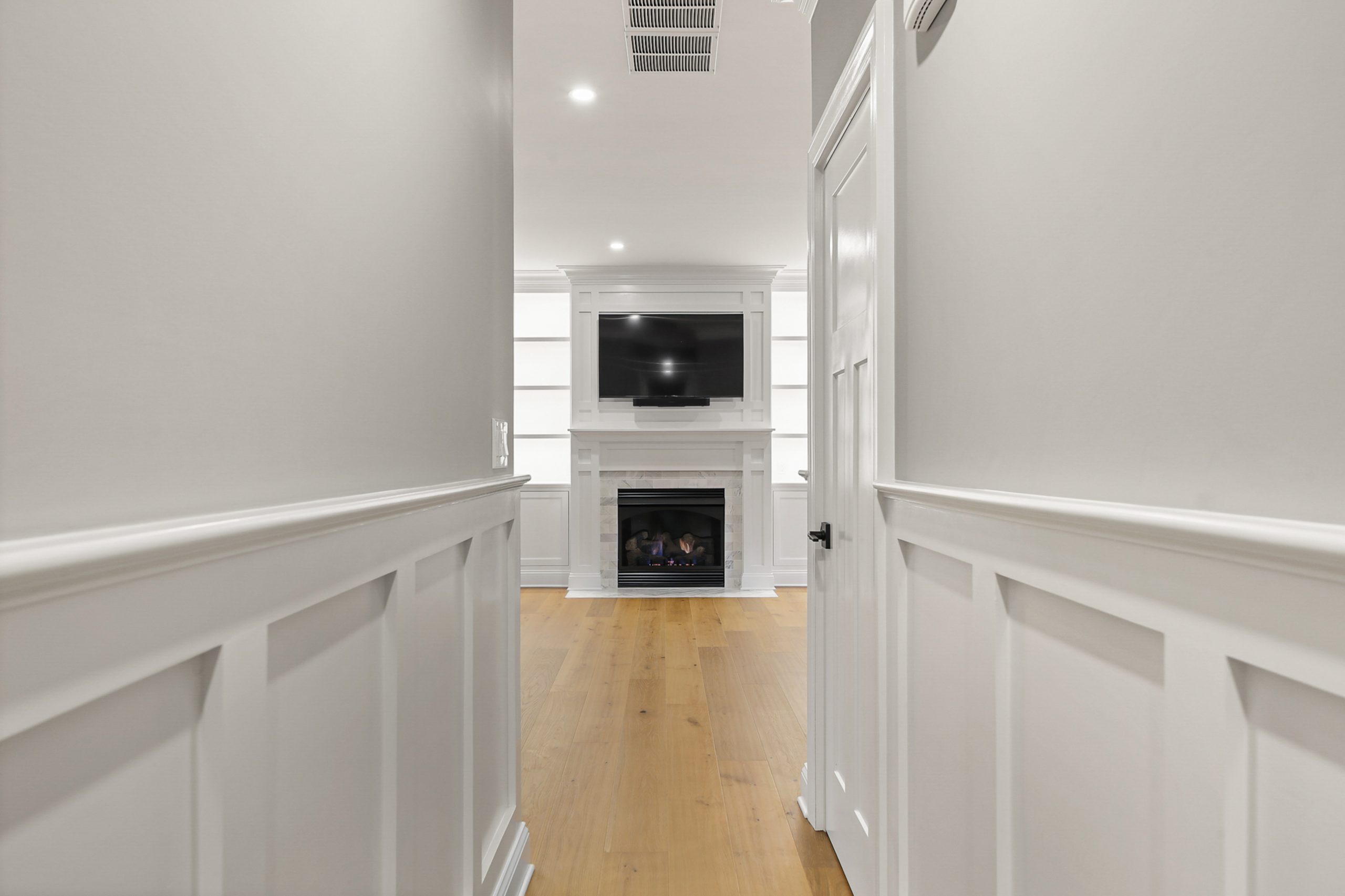 A hallway with white wainscoting and light wood floors leads to a living area featuring a built-in white fireplace, shelves, and a flat-screen TV mounted above the mantel.