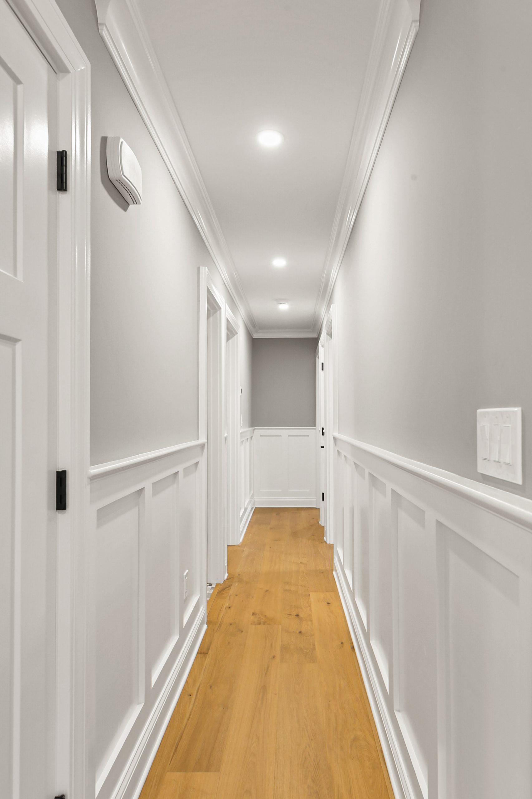 A bright hallway with light gray walls, white wainscoting and trim, recessed ceiling lights, and light wood flooring. Several closed white doors line the hallway.