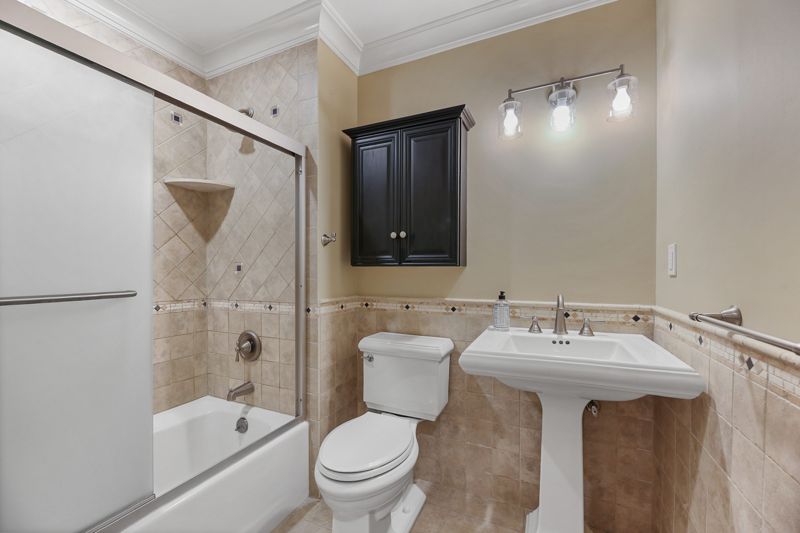 A traditional bathroom with beige tiled walls, a white bathtub with glass doors, a pedestal sink, a toilet, a black wall cabinet, and a three-bulb vanity light above the sink.