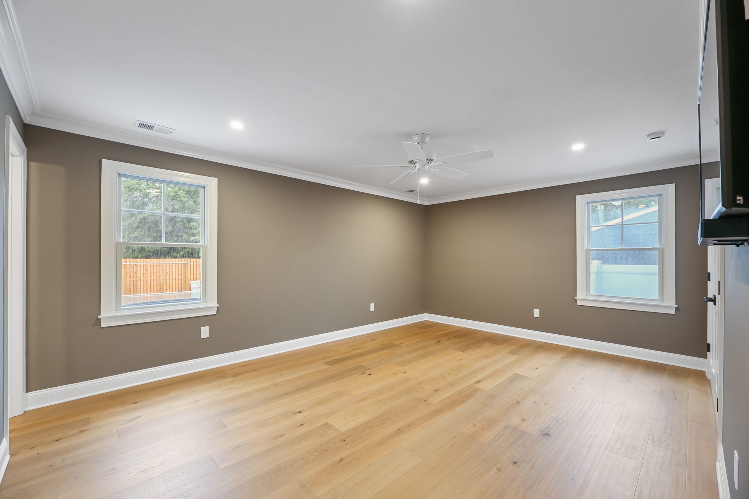 Spacious empty room with light wood flooring, taupe walls, white trim, two large windows, ceiling fan, and recessed lighting. Natural light streams in through the windows, highlighting the clean, modern look.
