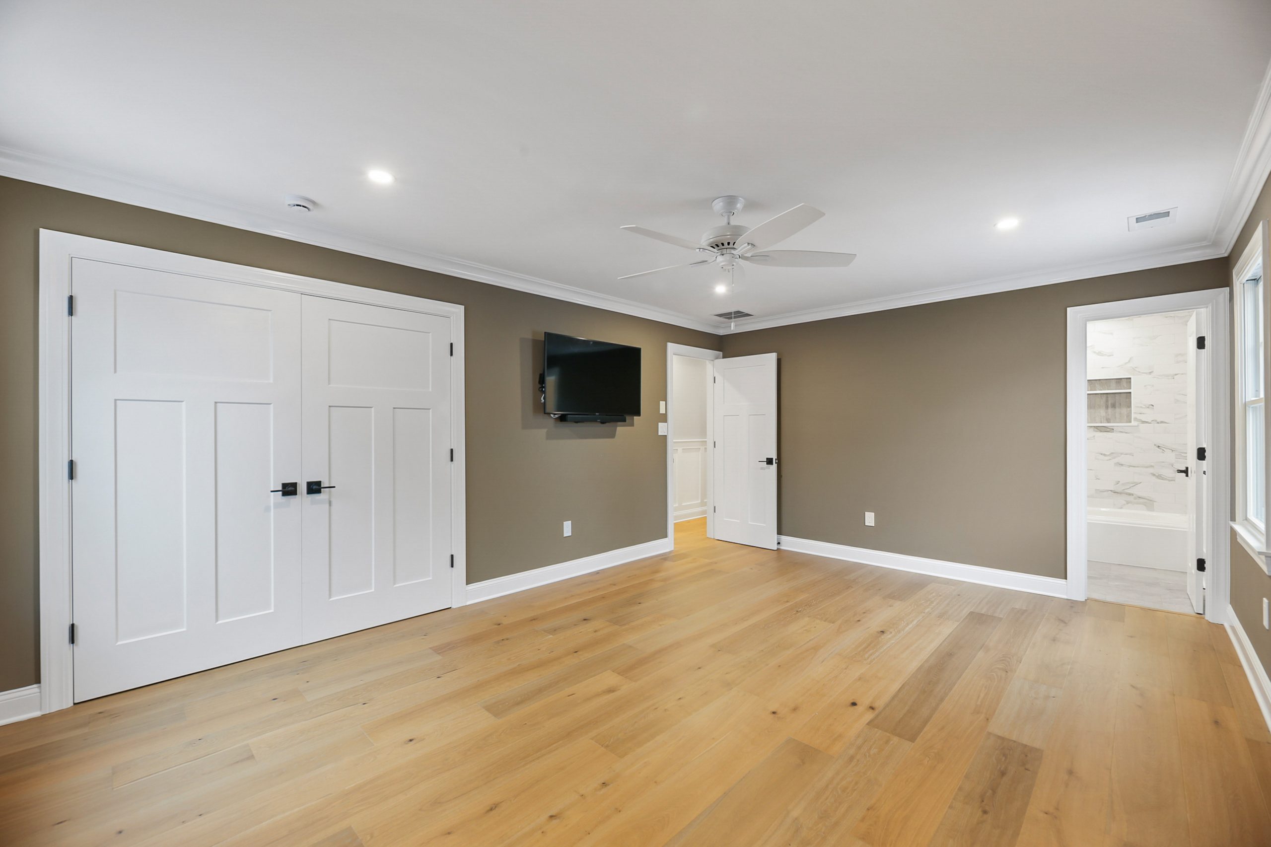 Spacious empty bedroom with light wood floors, taupe walls, white trim, double closet doors, a wall-mounted TV, ceiling fan, and a bathroom visible through an open door.
