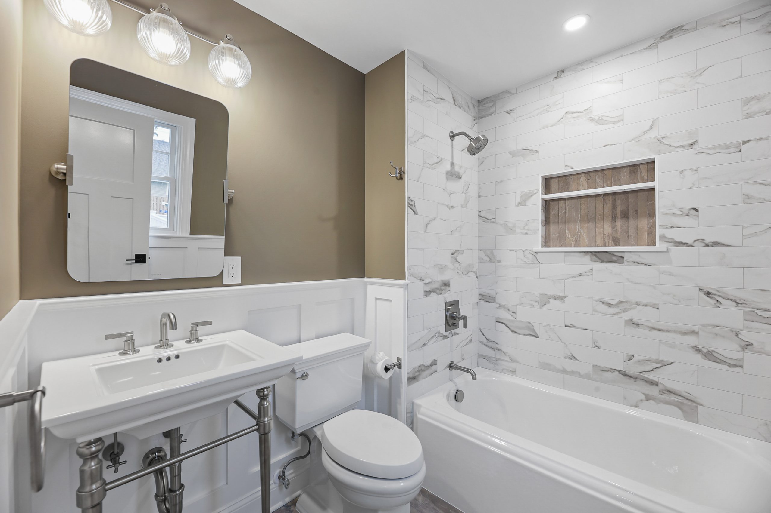 Modern bathroom with white and beige walls, a pedestal sink, toilet, and a bathtub with a marble-tiled shower. A large mirror is above the sink, and shelves are built into the shower wall.