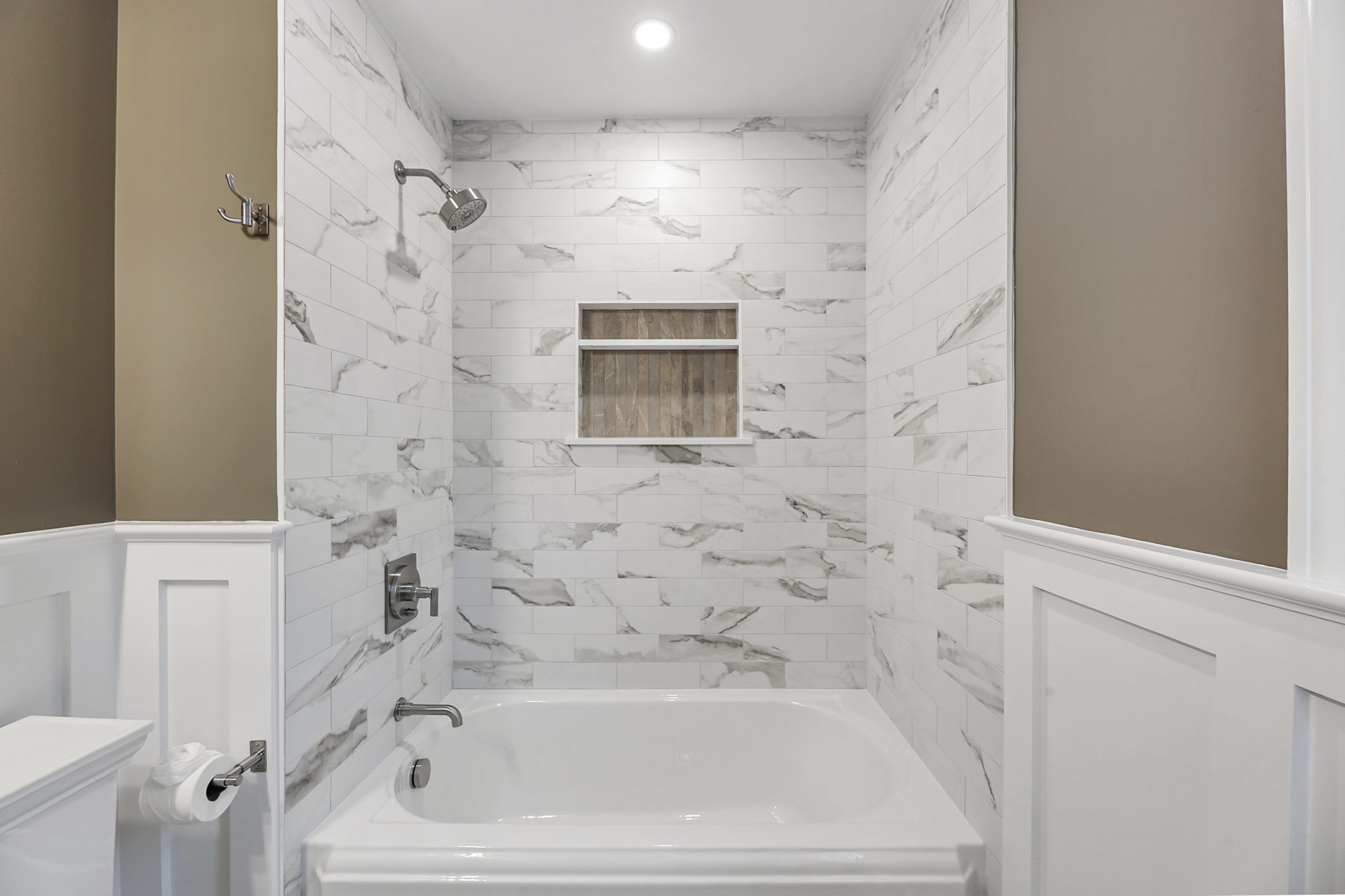 Modern bathroom with a white bathtub, marble-patterned tile walls, built-in recessed shelf, and silver showerhead and faucet. Walls feature white wainscoting and beige paint. Ceiling light above the shower area.