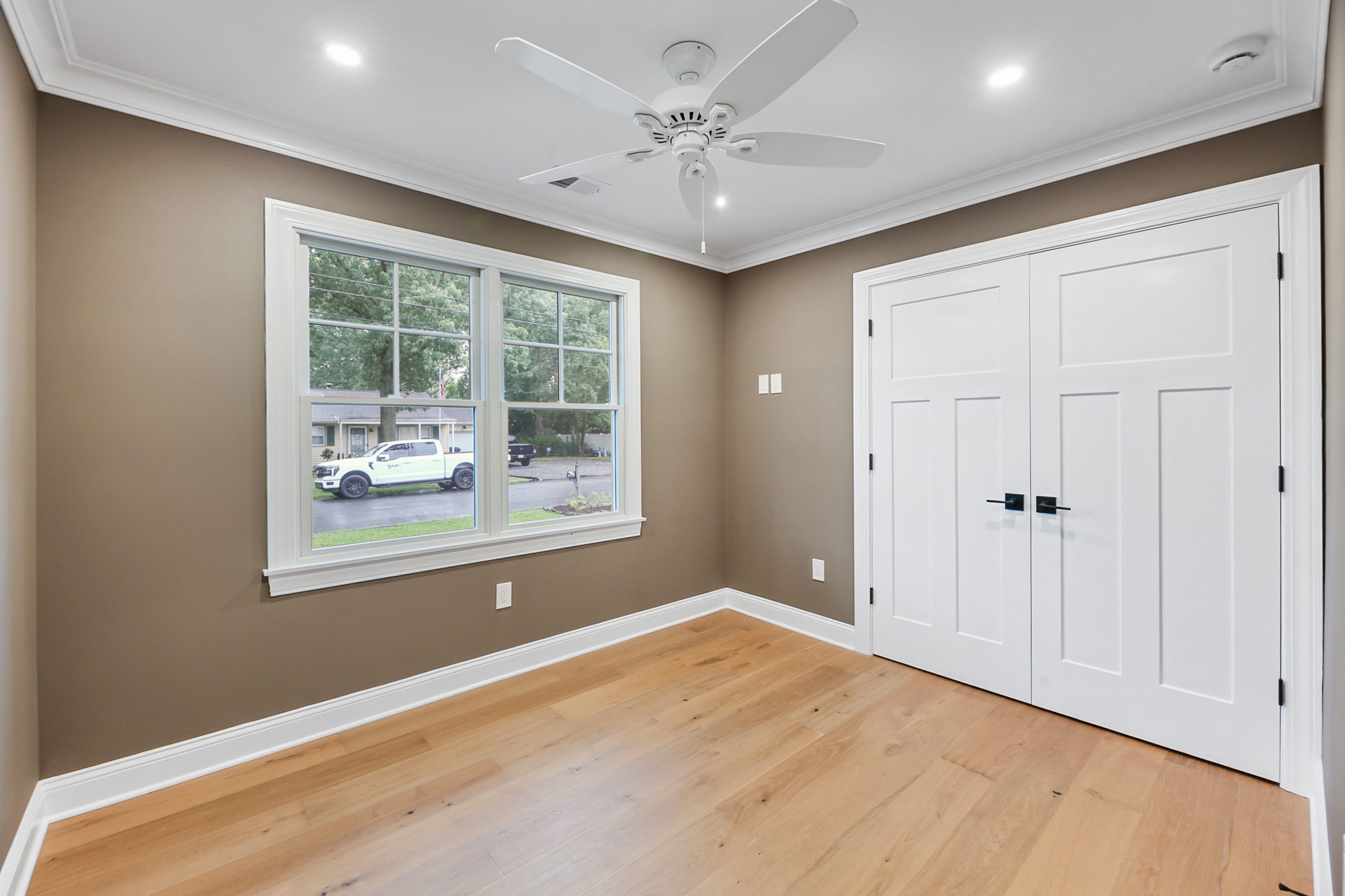 An empty room with beige walls, light wood flooring, a white ceiling fan, recessed lights, a large window showing cars and trees outside, and double white closet doors.