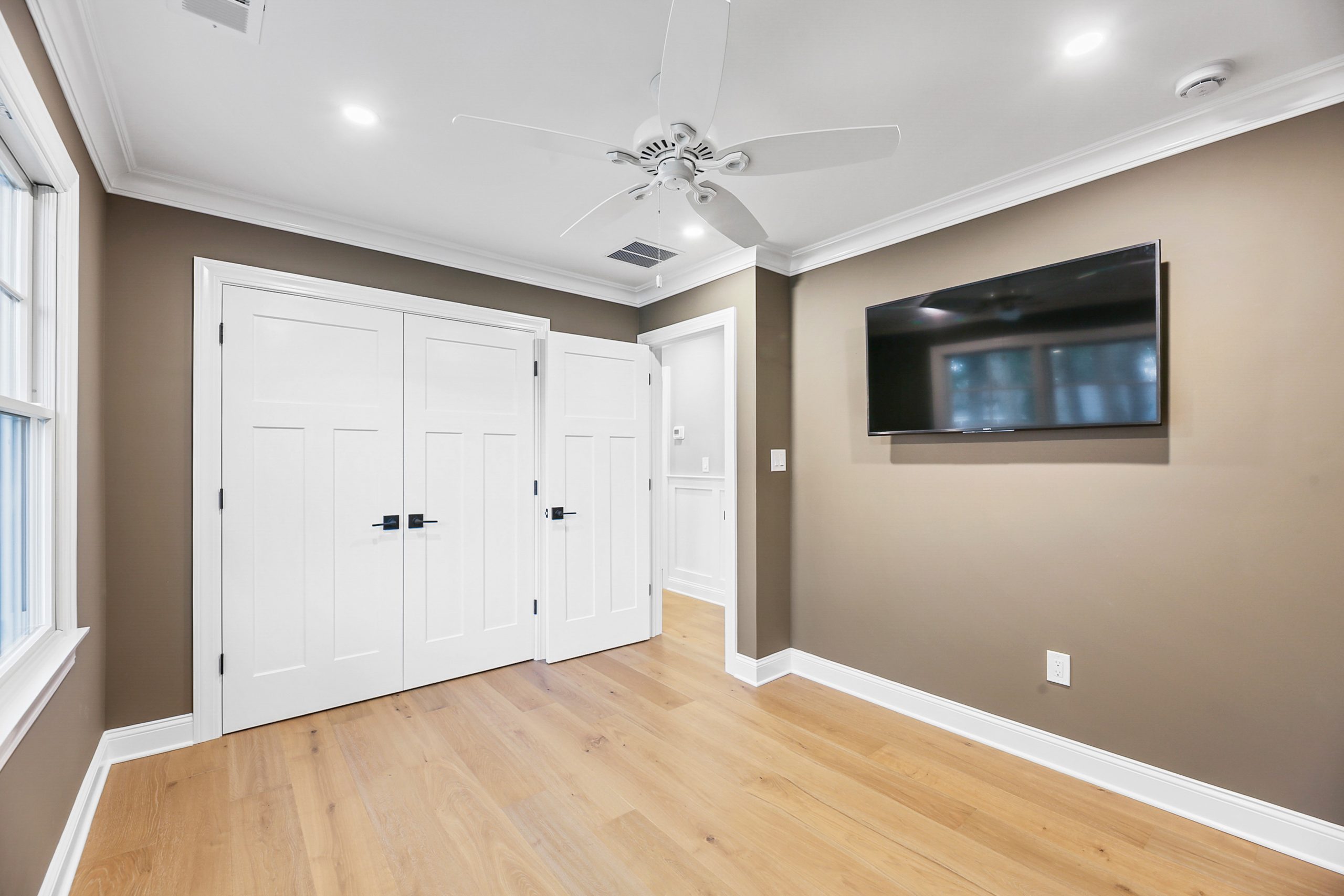 A modern, empty room with light wood flooring, taupe walls, white trim, a ceiling fan, a wall-mounted flat-screen TV, double white closet doors, and a large window letting in natural light.