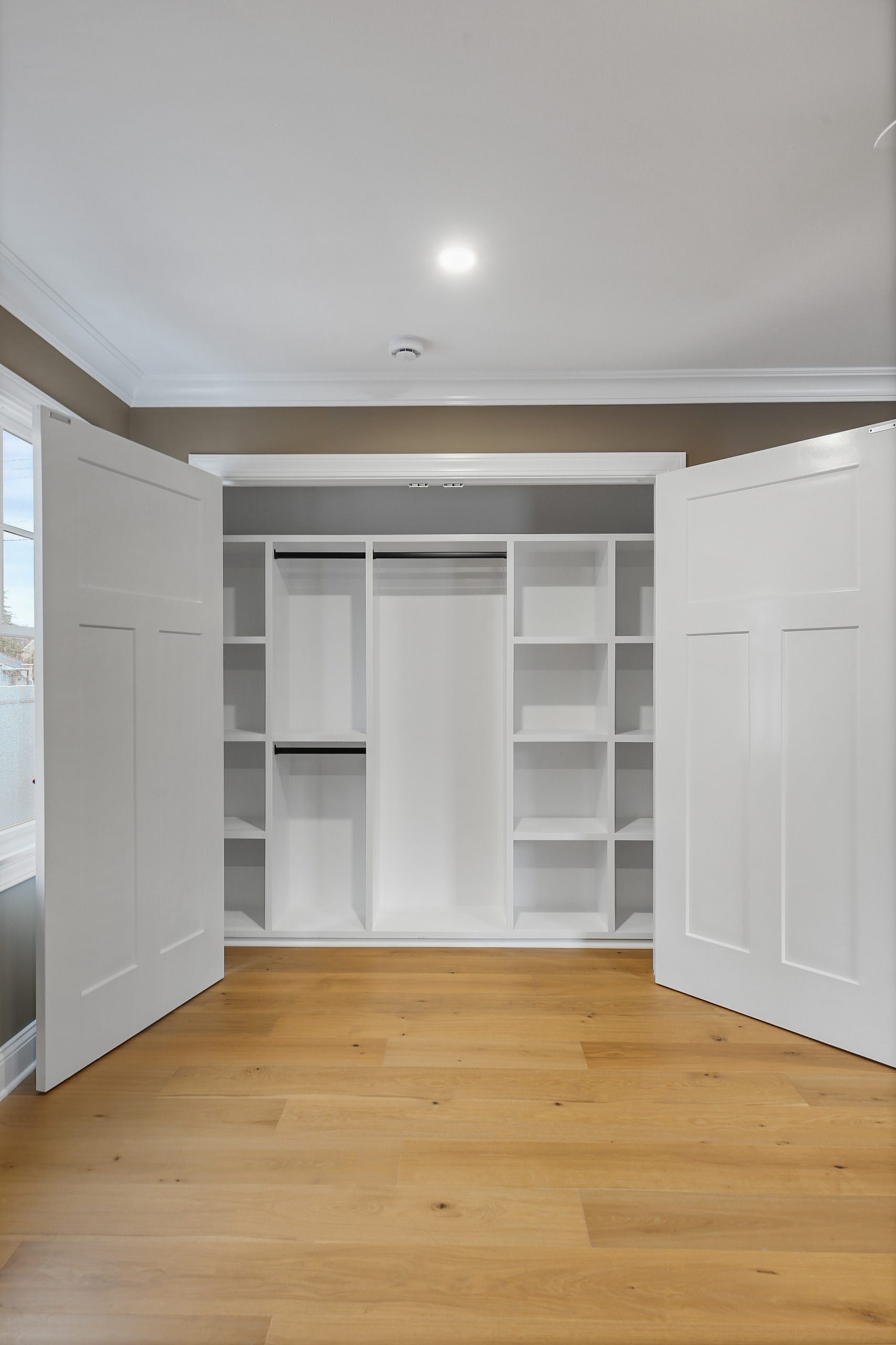 Open white double-door closet with built-in shelves and compartments, situated in a room with light wooden flooring and neutral-colored walls. Natural light enters from a window on the left.