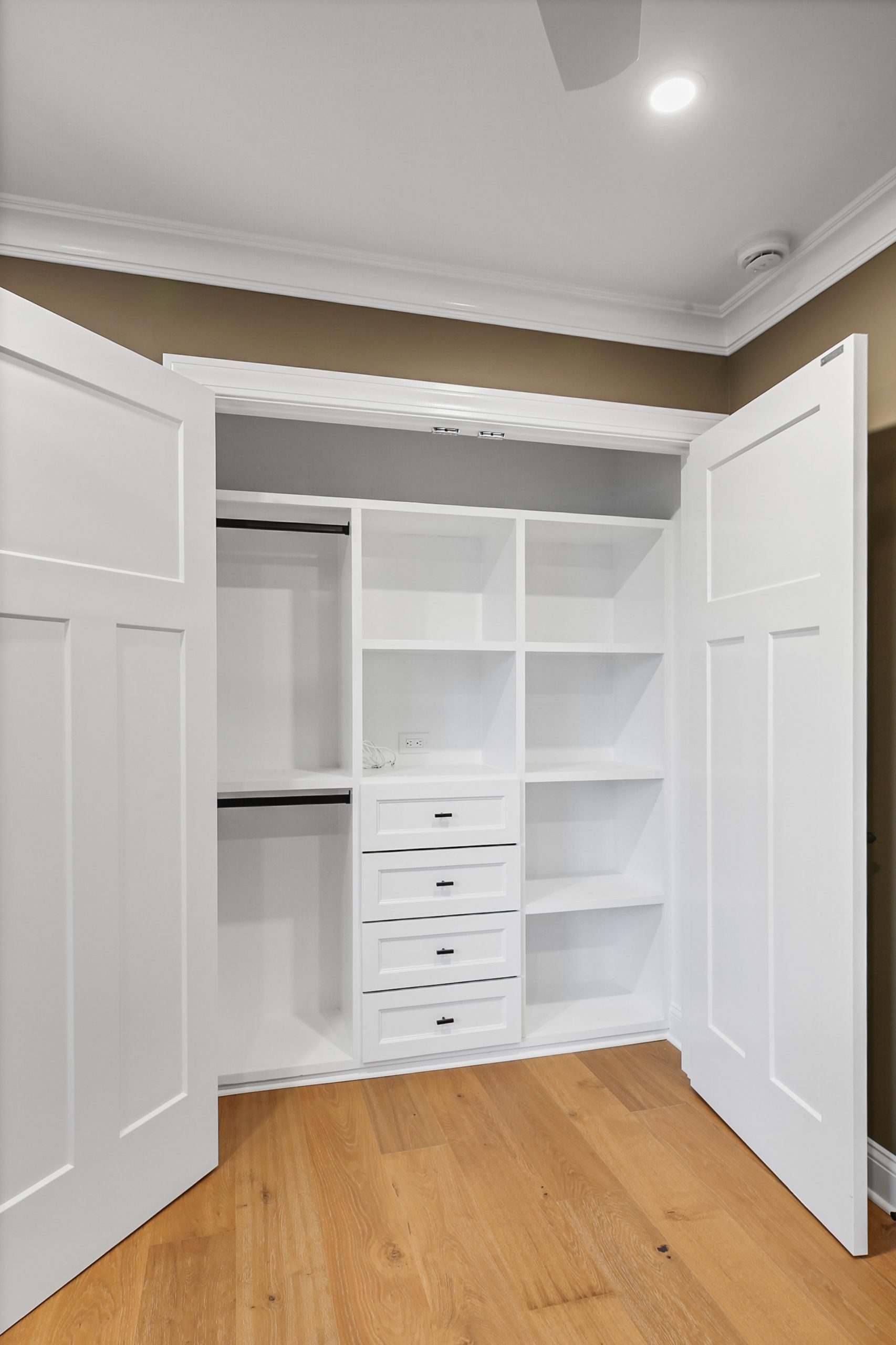 Open white closet with double doors, featuring multiple shelves, four drawers with silver handles, and two hanging rods. The closet is empty and set against a tan wall, with light wood flooring below.
