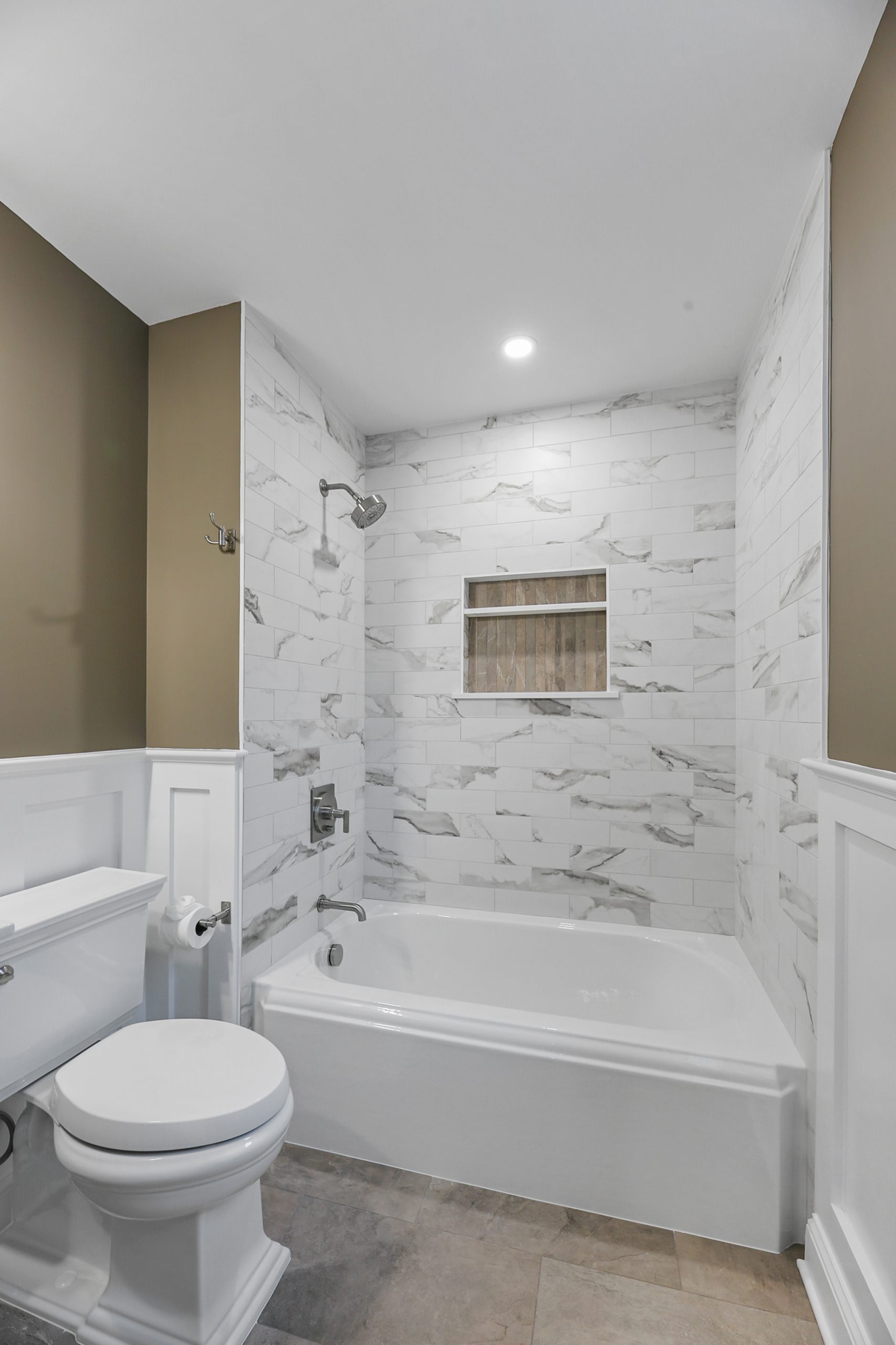 Modern bathroom with a white toilet, bathtub, and shower. The shower walls are tiled with white and gray marble, featuring a built-in shelf. The walls are painted olive green and white wainscoting is present.