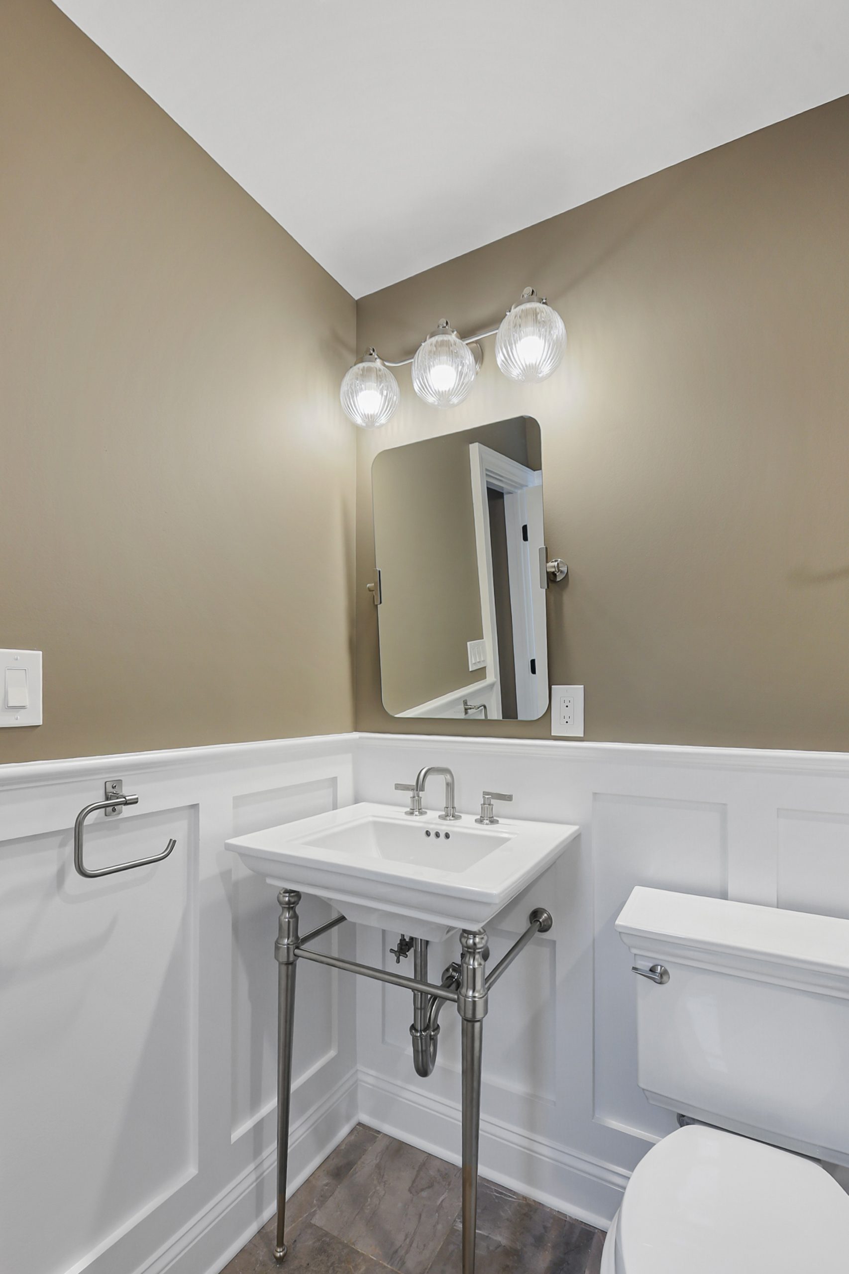 A small bathroom with a square sink, metal legs, wall-mounted faucet, rectangular mirror, three-globe vanity light, white wainscoting, taupe upper walls, a towel ring, and a white toilet.