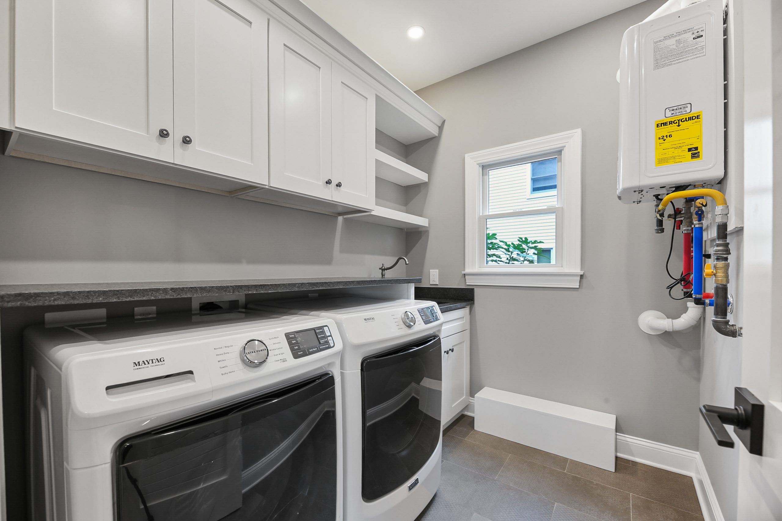 A modern laundry room with a side-by-side washer and dryer, white cabinets, open shelves, a small window, gray countertops, and a mounted water heater on the wall.