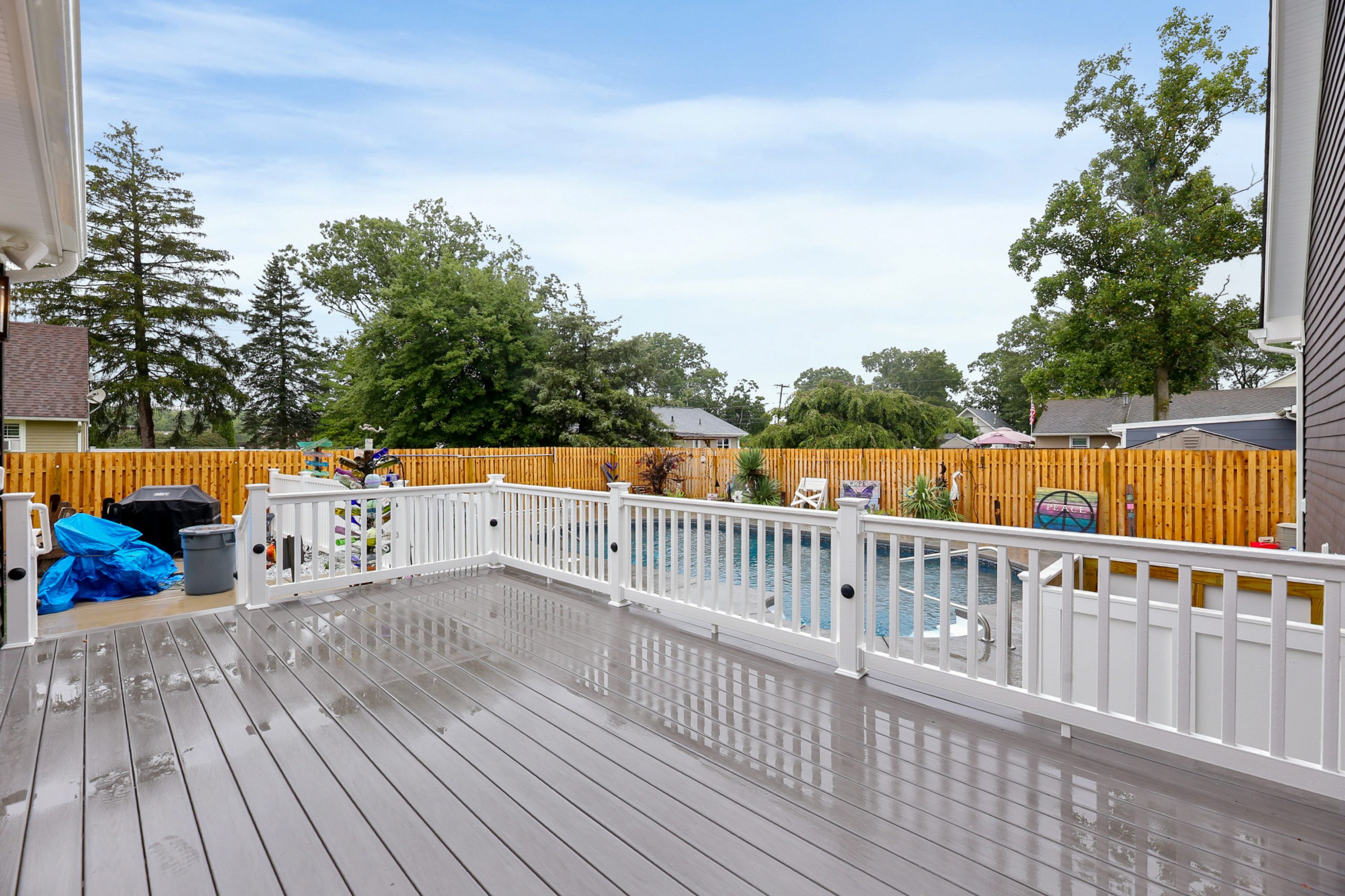 A spacious backyard deck with white railings overlooks a swimming pool, surrounded by a wooden fence and trees. Patio chairs and pool toys are visible near the pool under a cloudy sky.