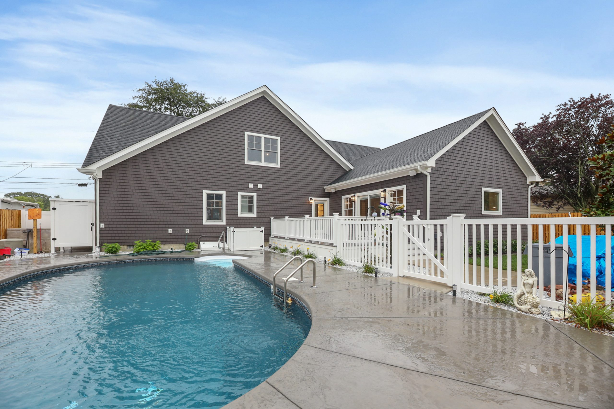 A backyard with a modern house, a curved swimming pool, wet concrete patio, white fence, and some greenery. The sky is partly cloudy.